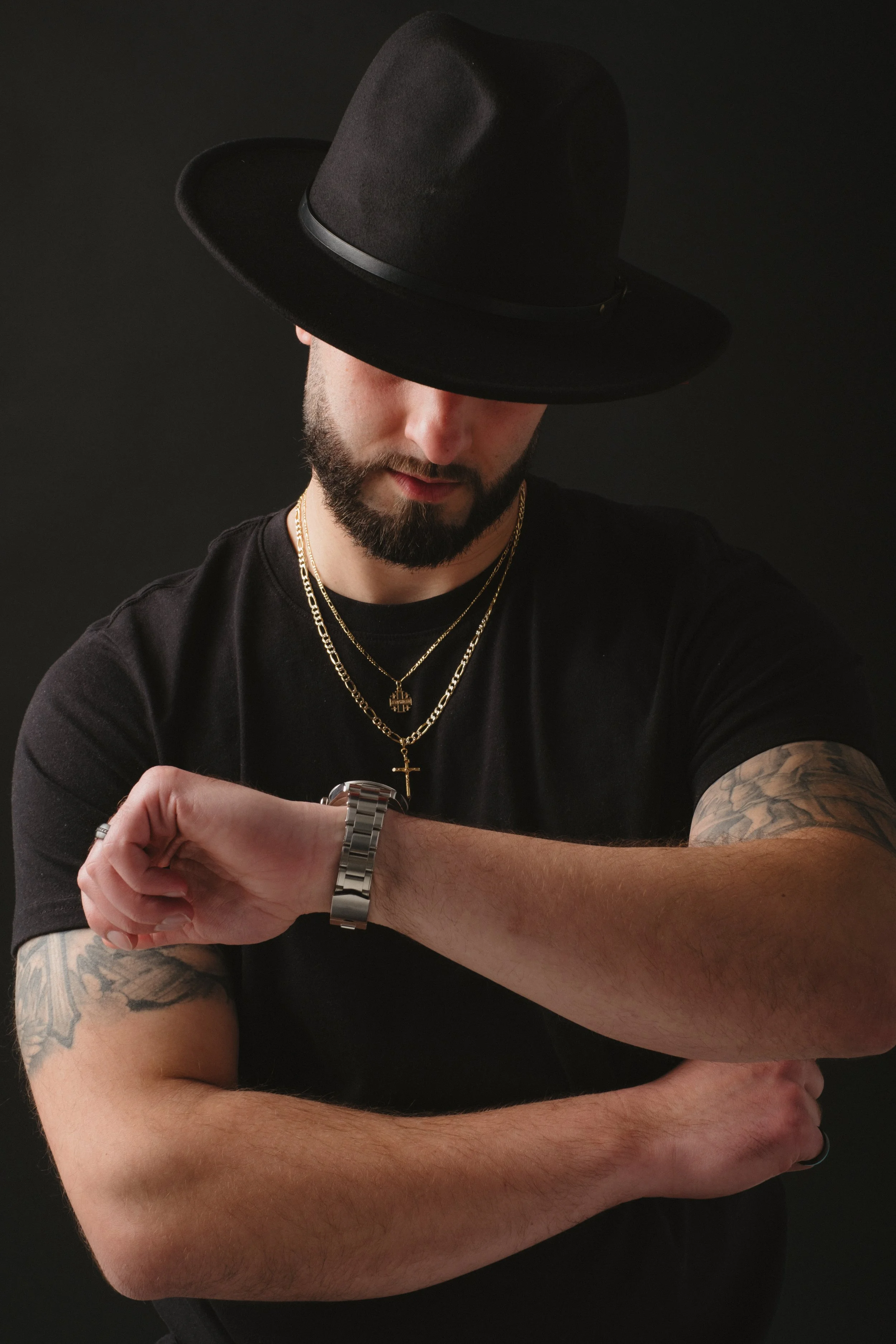 A man wearing a black hat, black t-shirt, gold chains, and a watch looks at his wrist with a black background.