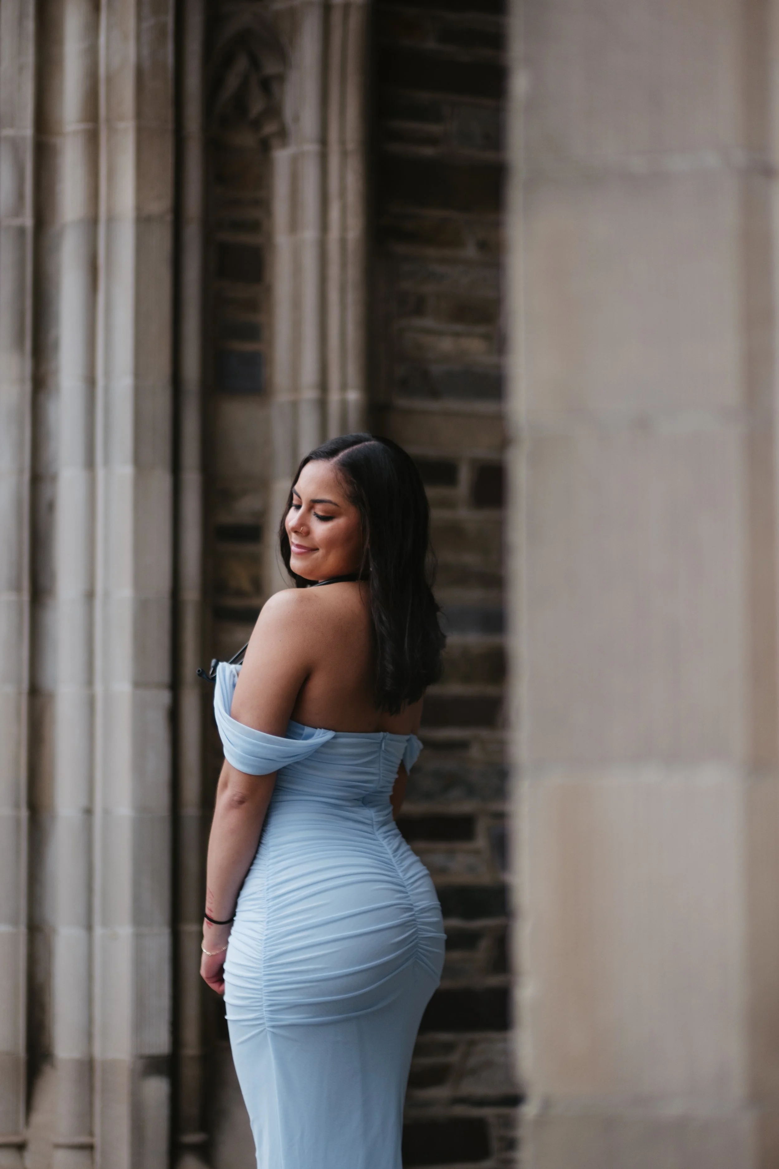 A woman in a light blue dress with back ruching, standing among stone columns in a historical building, smiling with eyes closed.