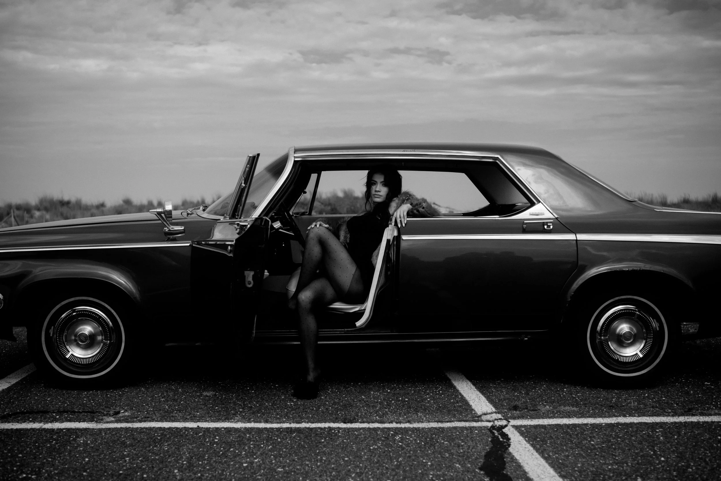 A woman sitting in the open doorway of a vintage car parked on a road, with a landscape background and cloudy sky, in black and white.