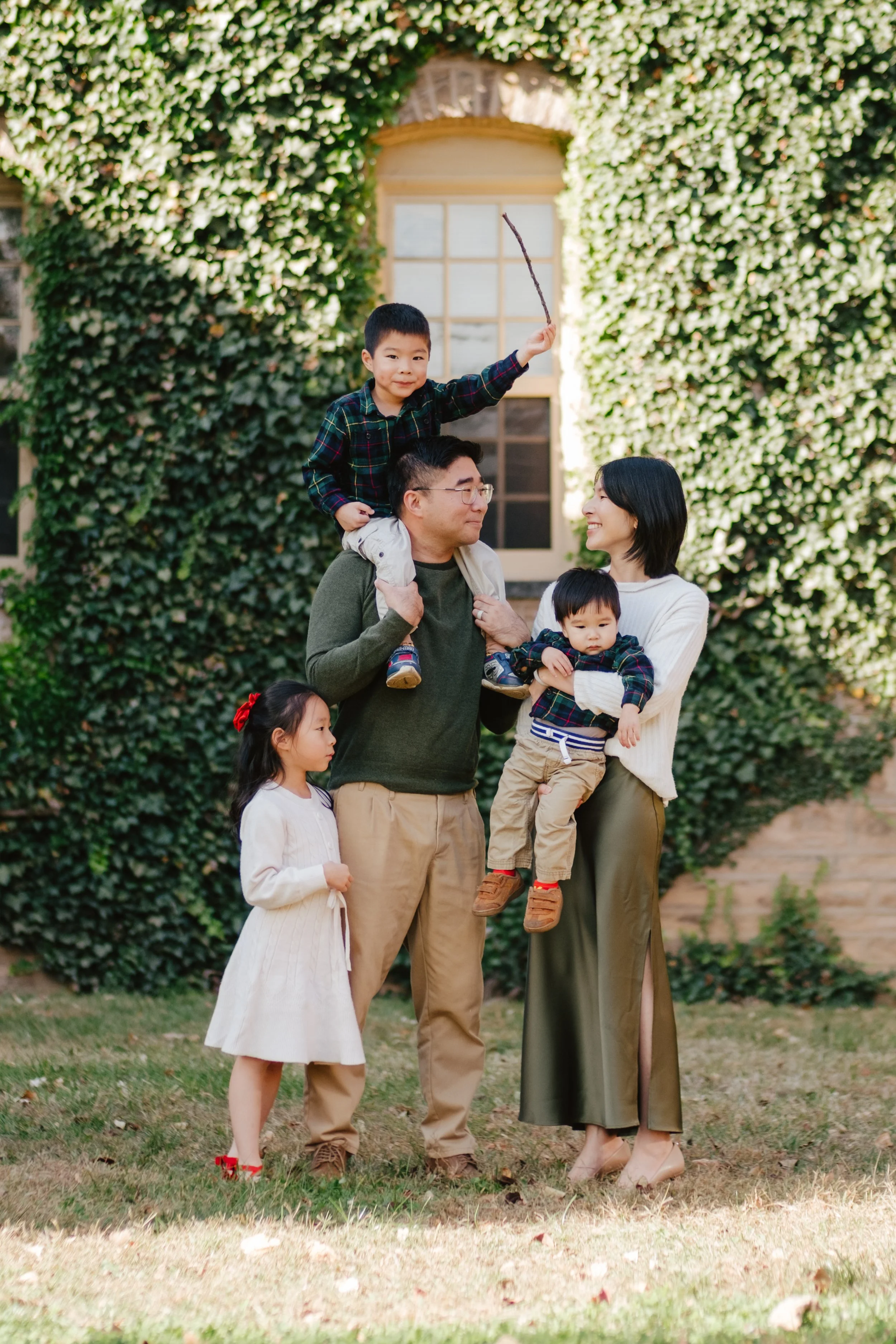 A happy family of five outdoors in front of a countryside stone house with green ivy. The father carries a boy on his shoulders, while the mother holds a younger boy. A girl stands beside the father looking at the family.