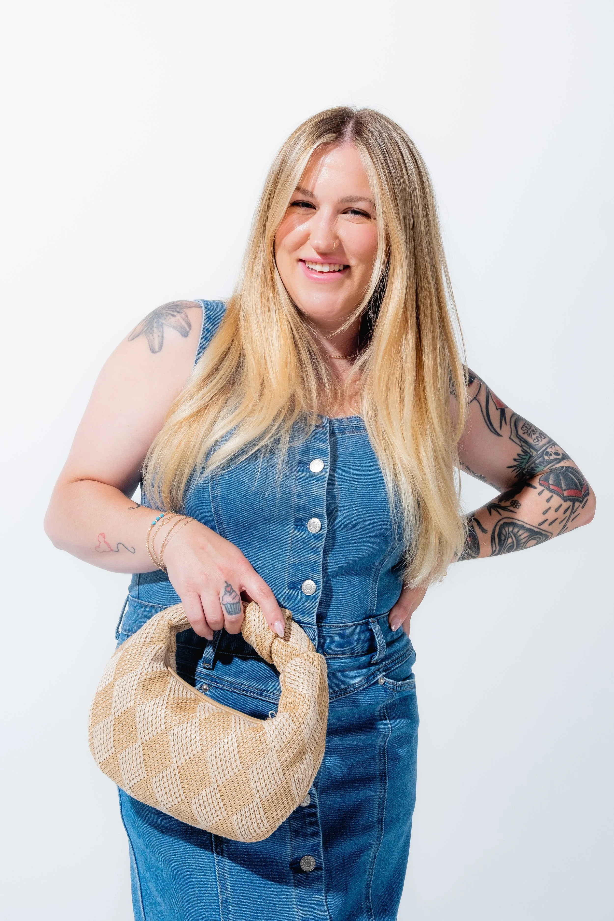 A woman with long blonde hair, tattoos on her arms, and a nose ring, wearing a sleeveless denim dress, holding a beige woven handbag, smiling and posing against a plain white background.