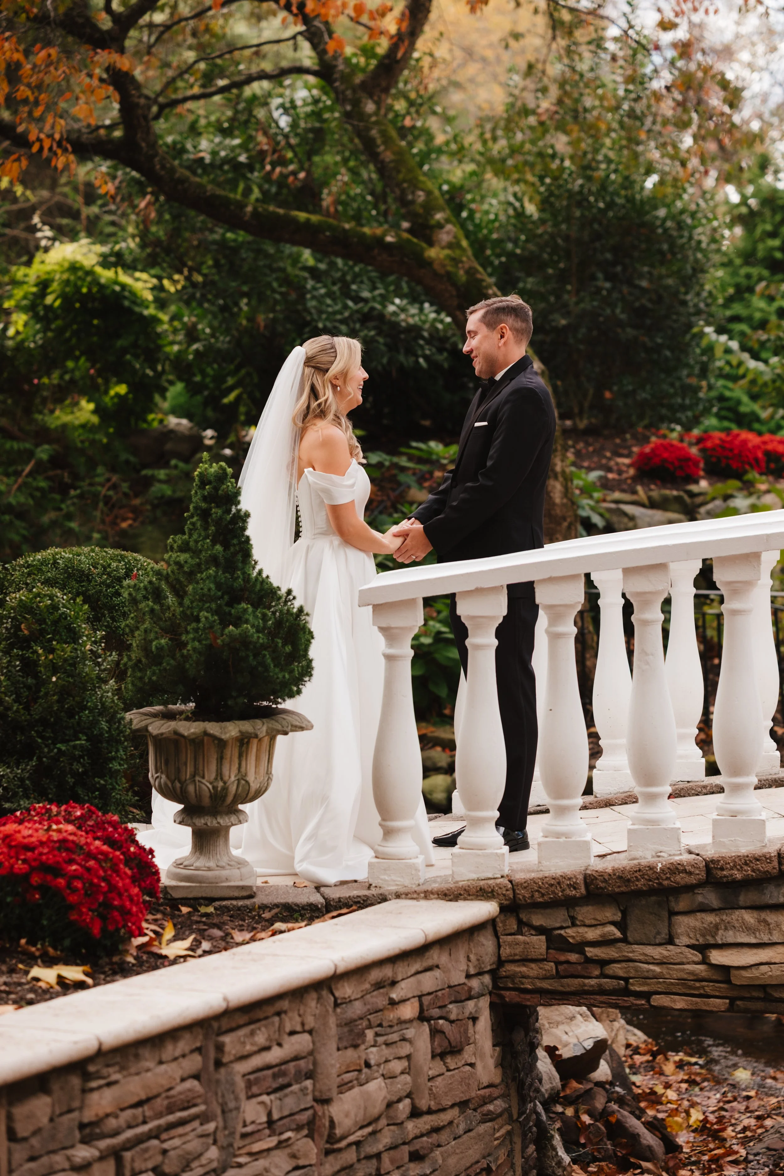 A bride and groom holding hands and smiling at each other on a white bridge in a garden during autumn.