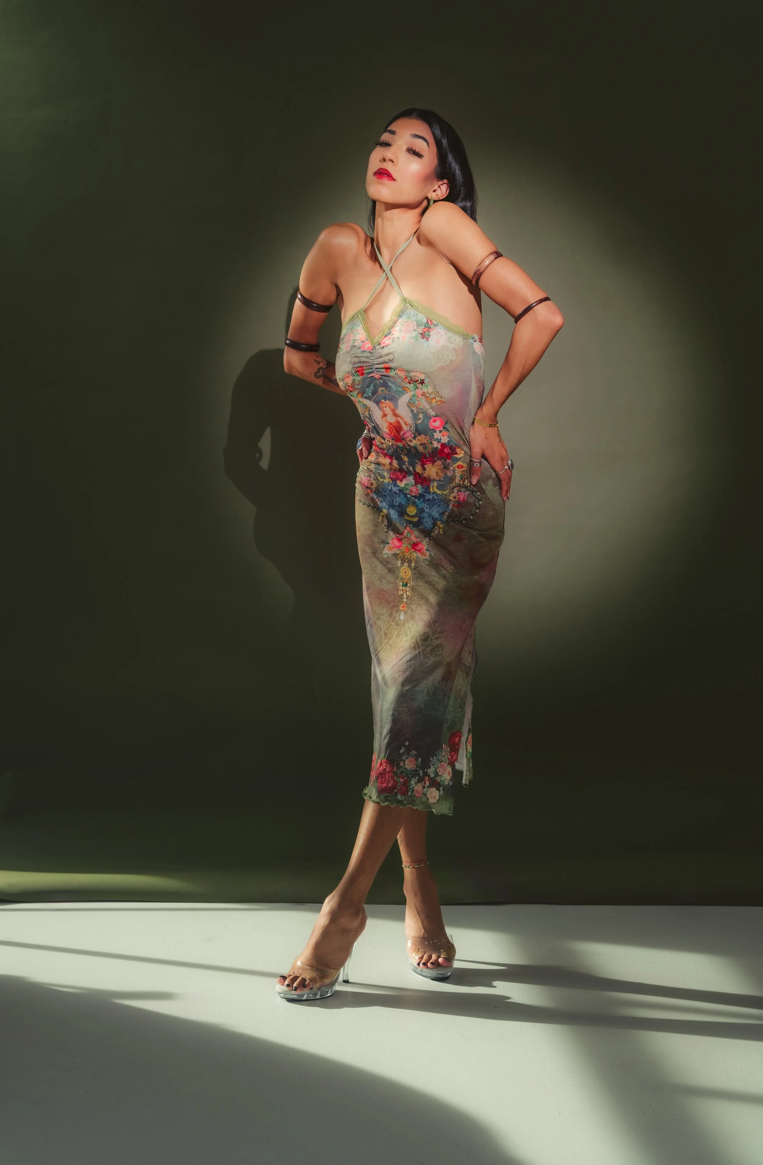 A woman with dark hair and red lipstick wearing a sleeveless, embroidered dress and transparent high heels, posing against a dark background.
