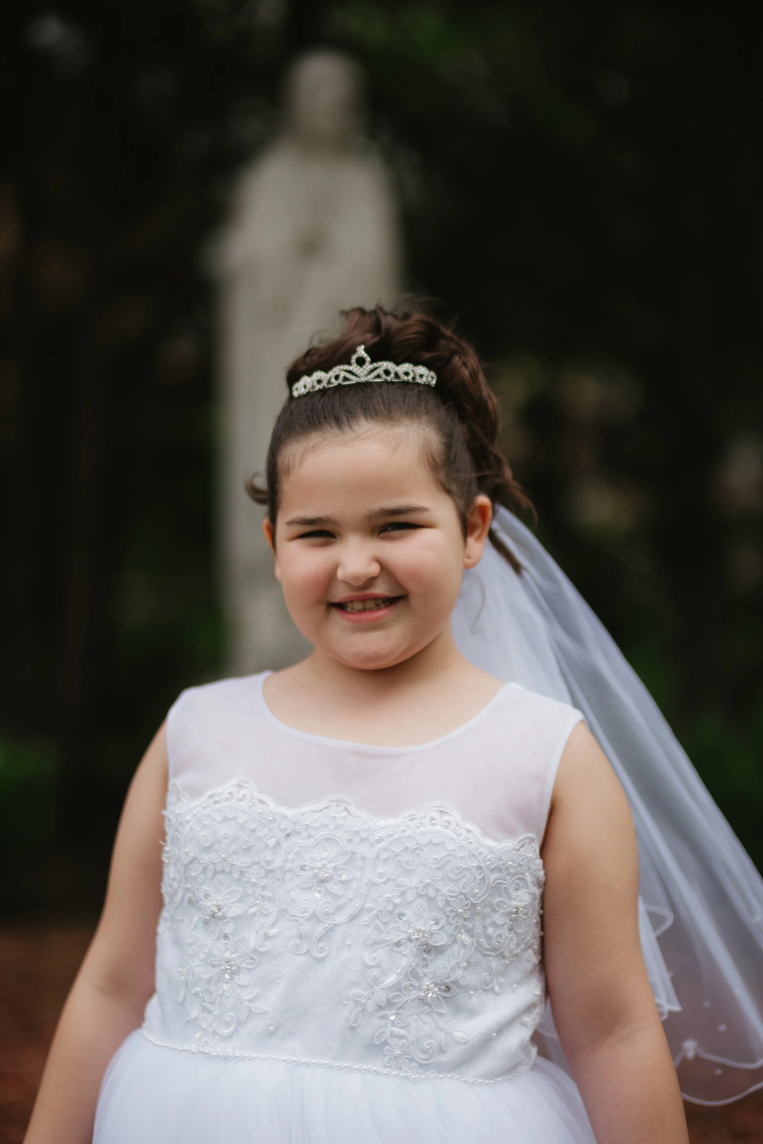 Young girl in a white dress with lace embroidery, wearing a tiara and veil, smiling outdoors with blurred background.