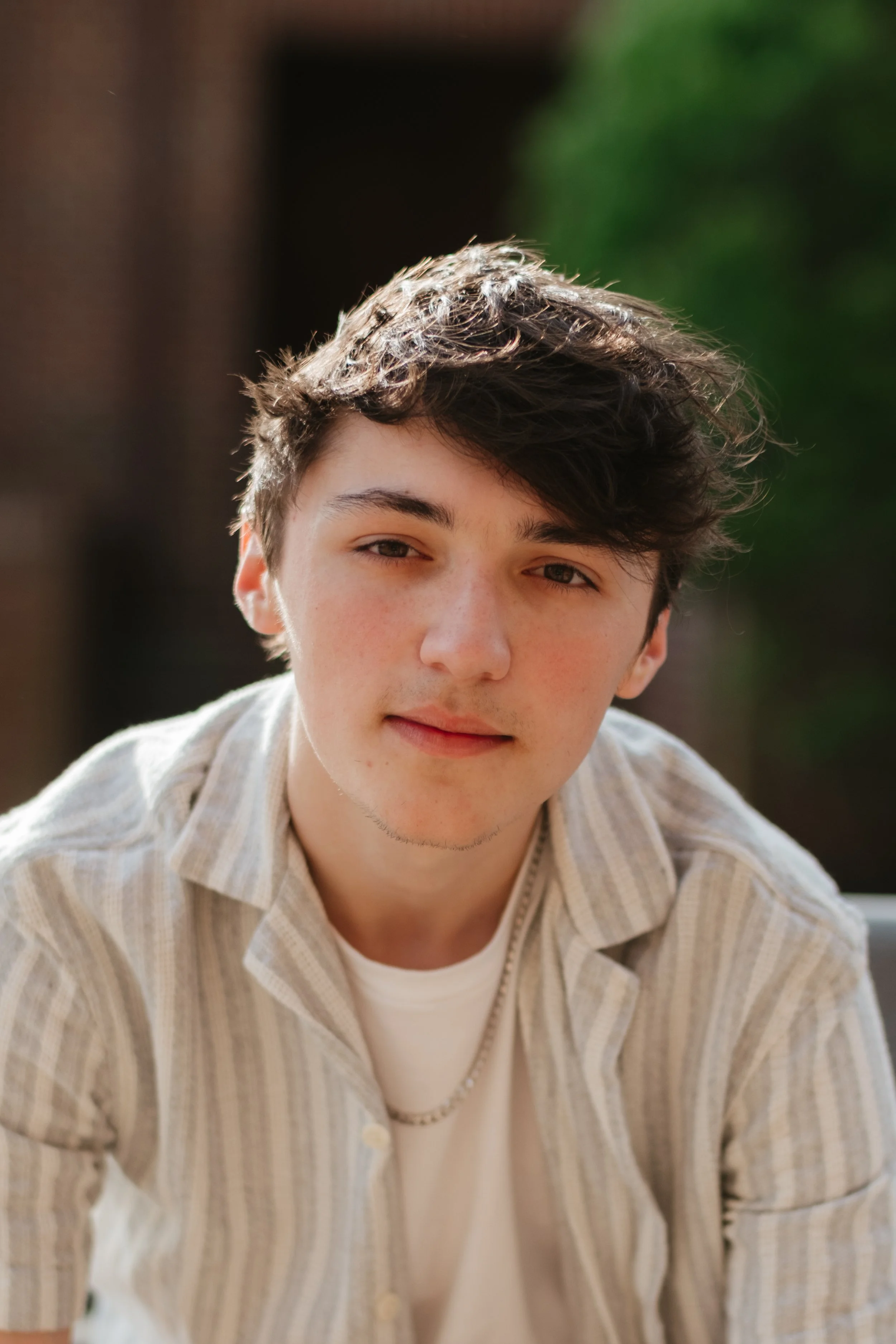 A young man with tousled dark hair, fair skin, and subtle facial hair, wearing a light-colored, striped button-up shirt over a white t-shirt, looking into the camera with a relaxed expression outdoors.