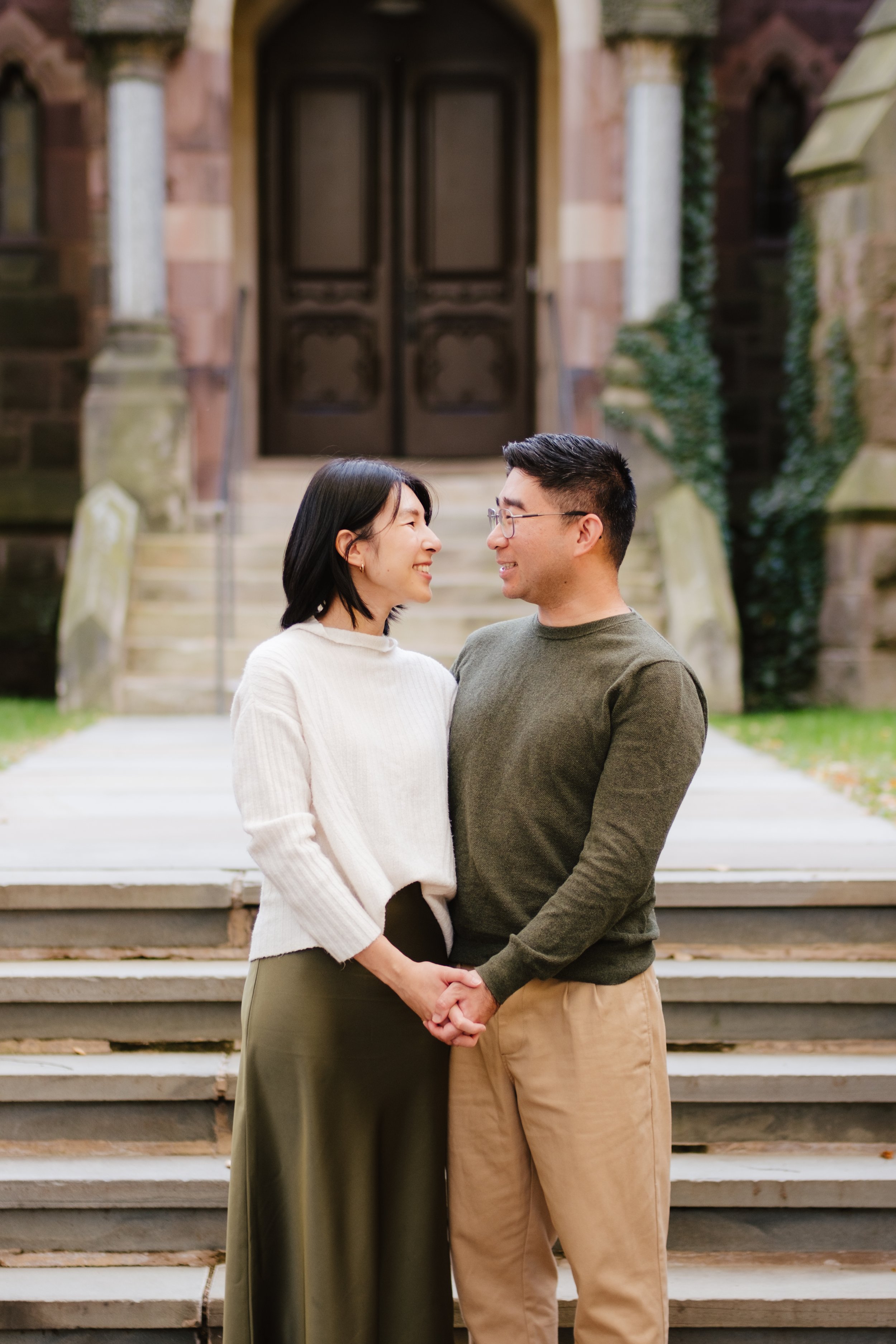 A happy couple holding hands and looking at each other in front of a historic building with stairs and large wooden doors.