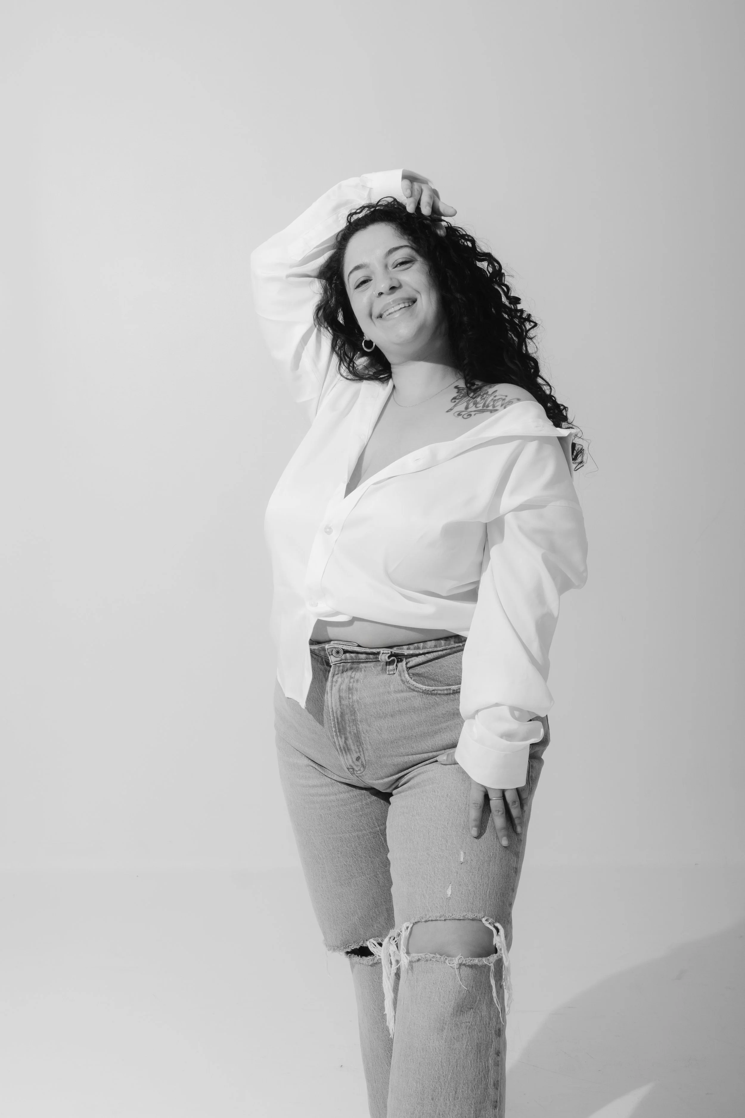 Black and white photo of a woman with curly hair smiling, wearing a white unbuttoned shirt and ripped jeans, standing against a plain background.