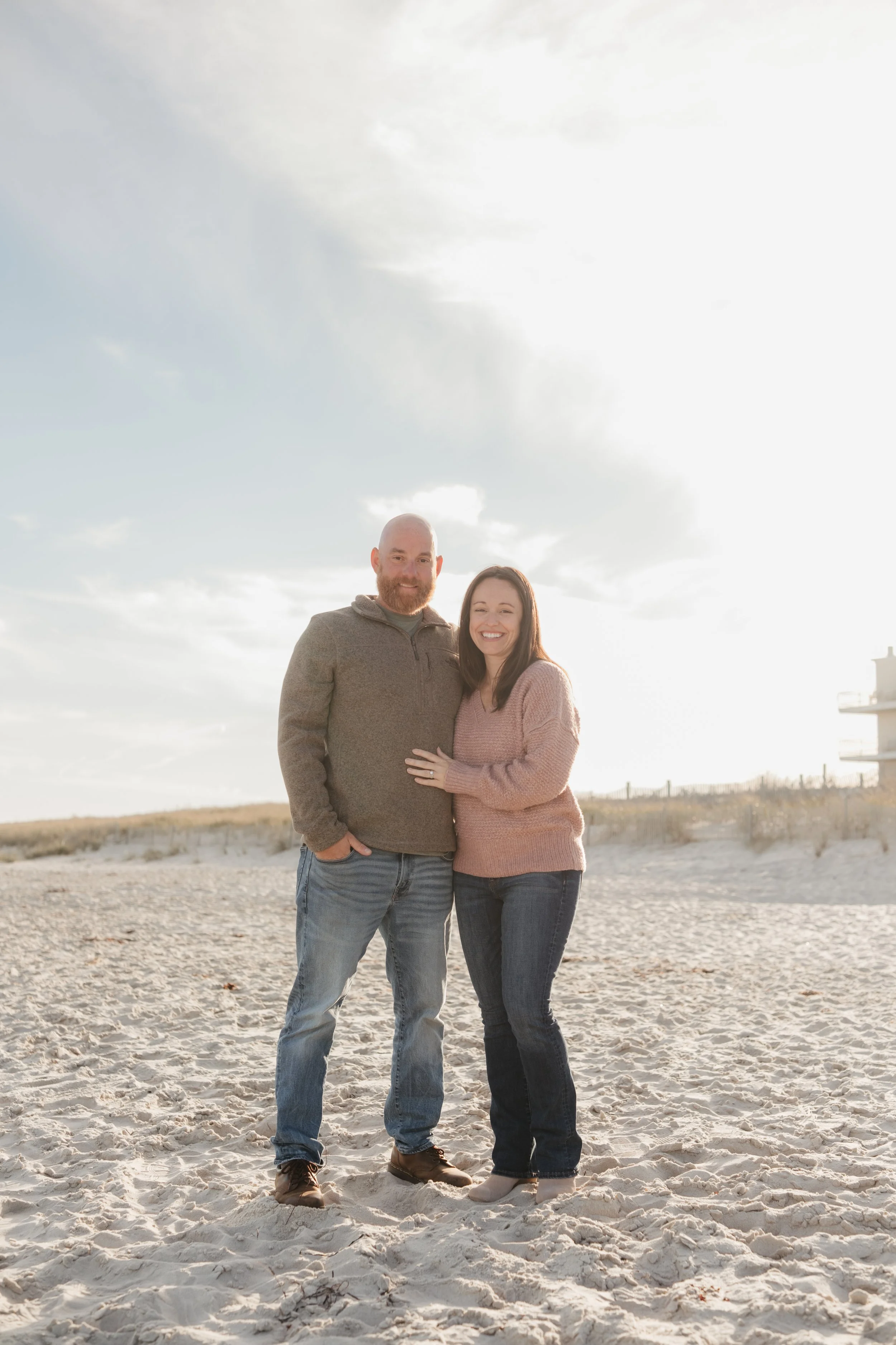 A smiling couple stands on a sandy beach, with the man in a gray sweater and jeans, and the woman in a pink sweater and jeans, during sunset.