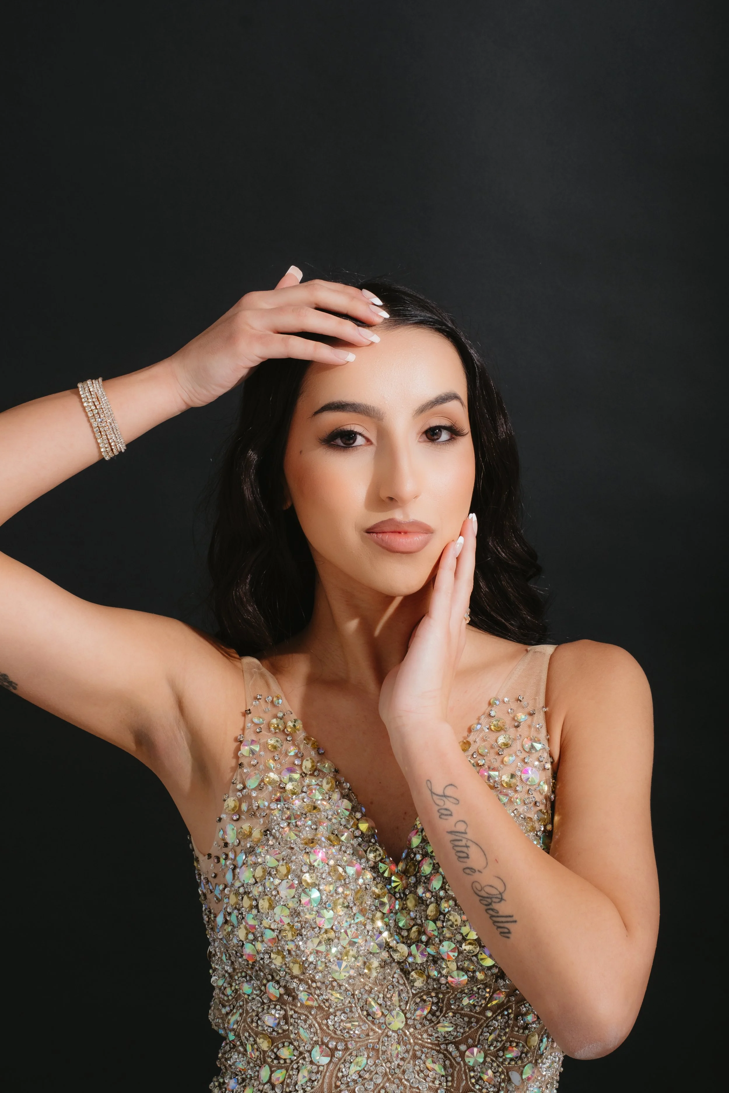 Portrait of a woman with black hair and tattoos, wearing a sleeveless dress with rhinestones, against a dark background.