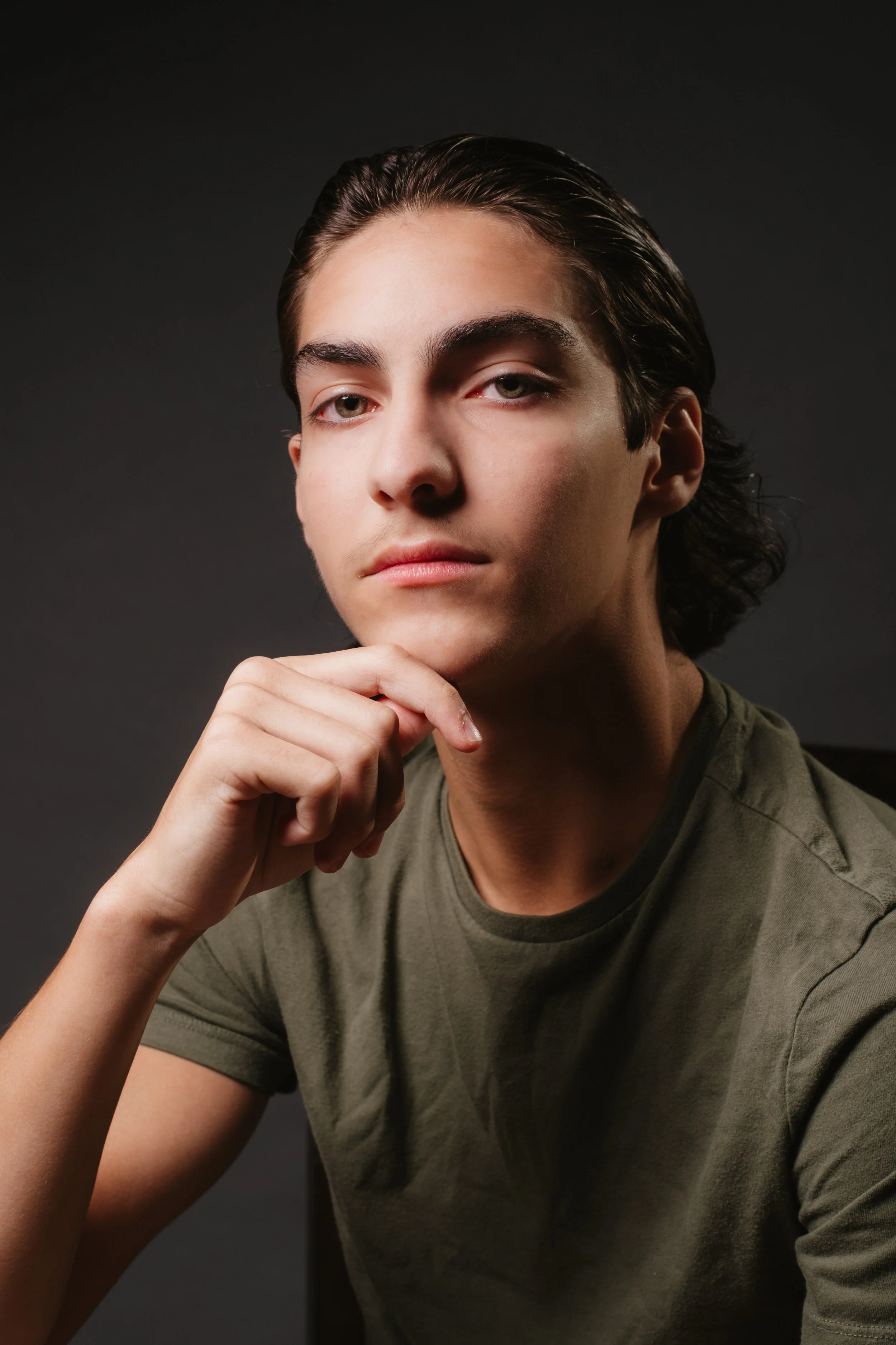 Portrait of a young man with dark hair, wearing a green t-shirt, sitting against a dark background, looking thoughtfully at the camera.