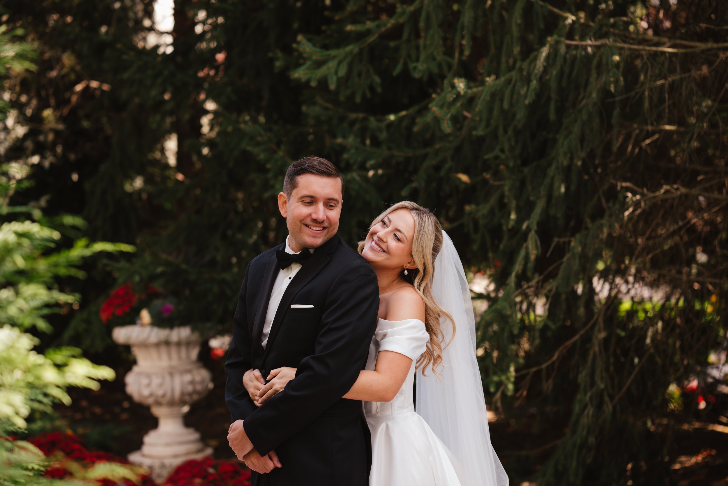 Bride and groom standing outdoors, smiling, with a background of green trees and red flowers.