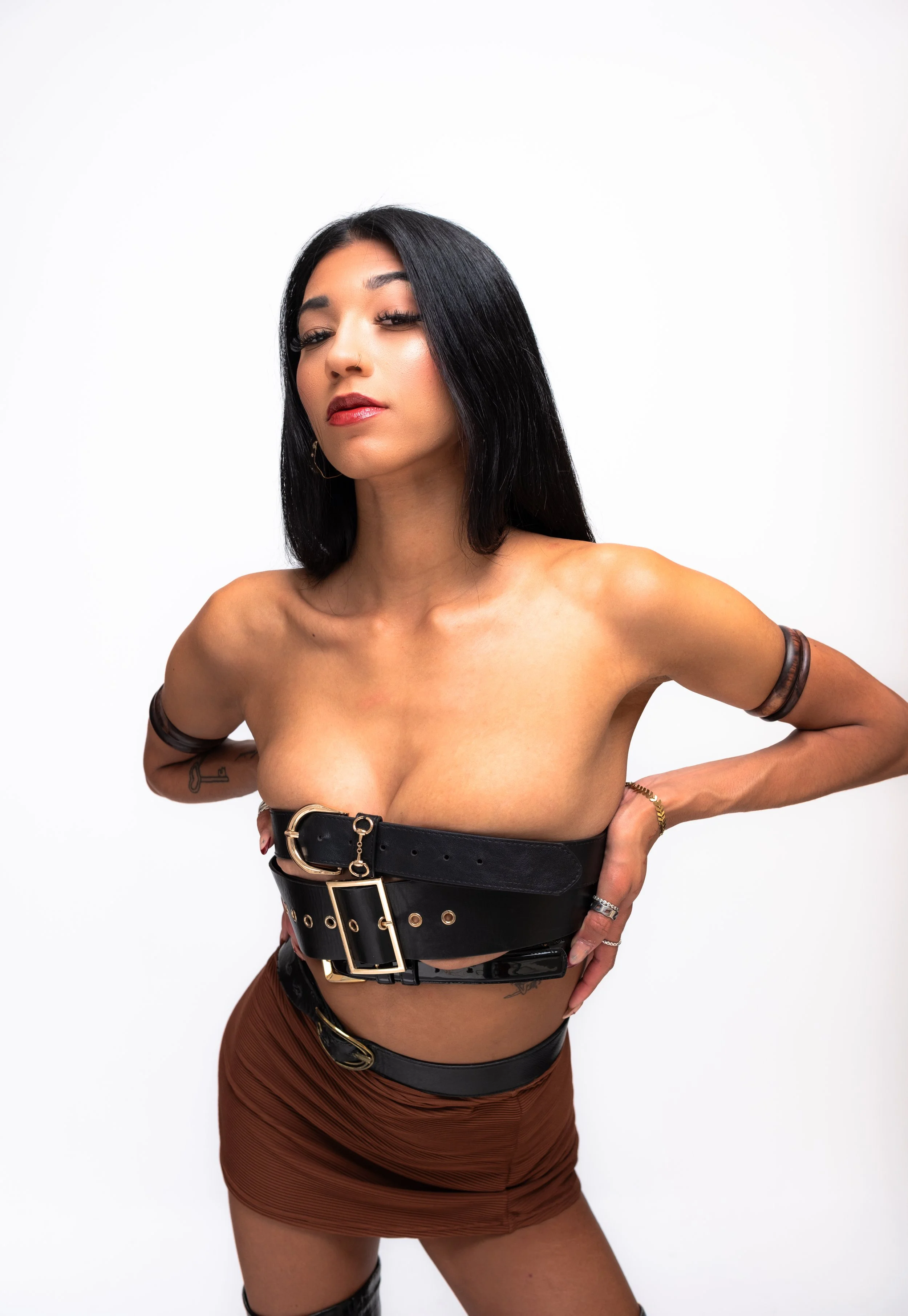 A woman with long black hair, dark eyeshadow, and red lipstick poses with her hands on her hips against a plain white background. She is wearing a black leather belt with gold accents around her chest and a brown skirt.