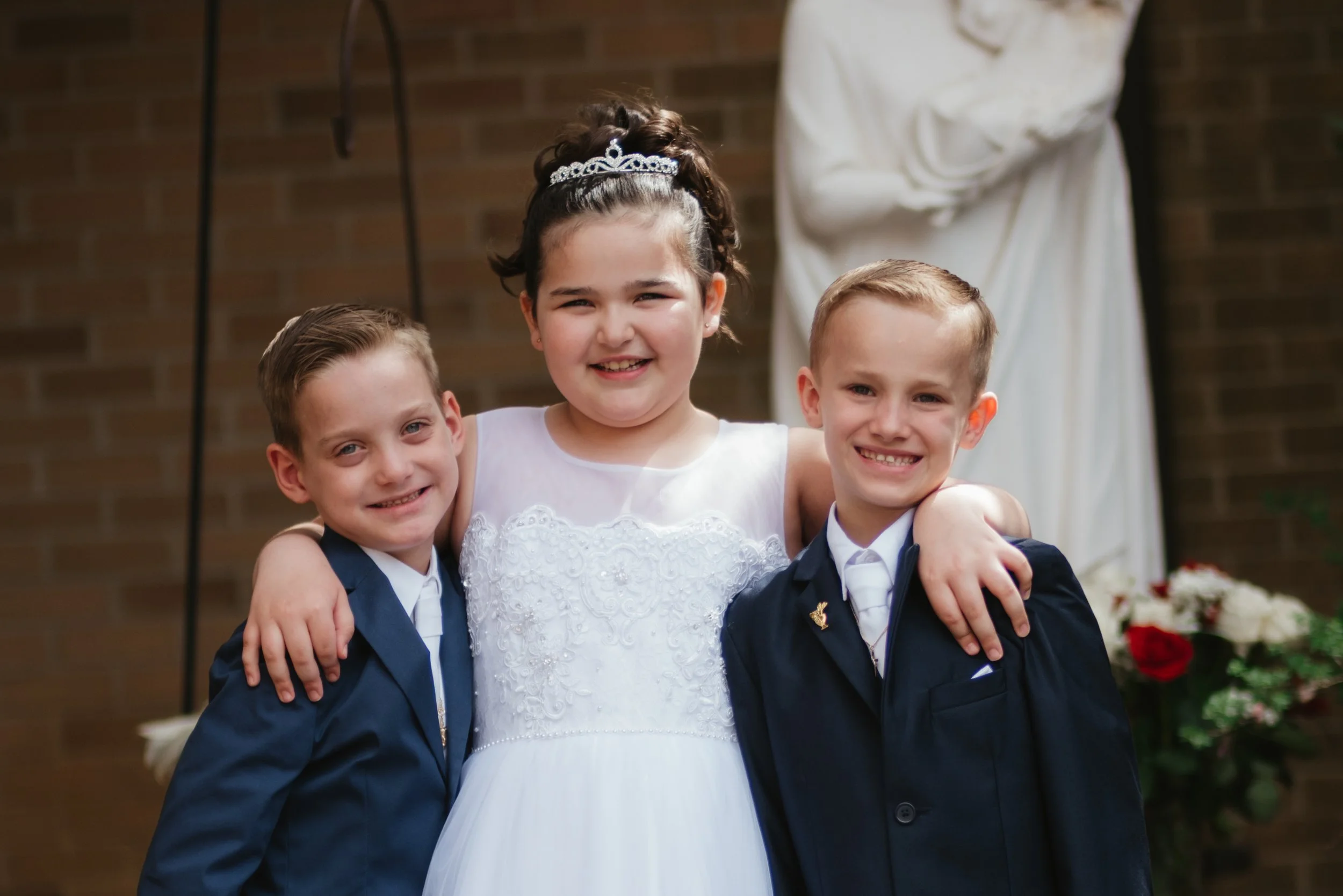 Three children in formal attire, a young girl in a white dress and tiara, and two boys in navy suits, smiling and posing together at a wedding ceremony.