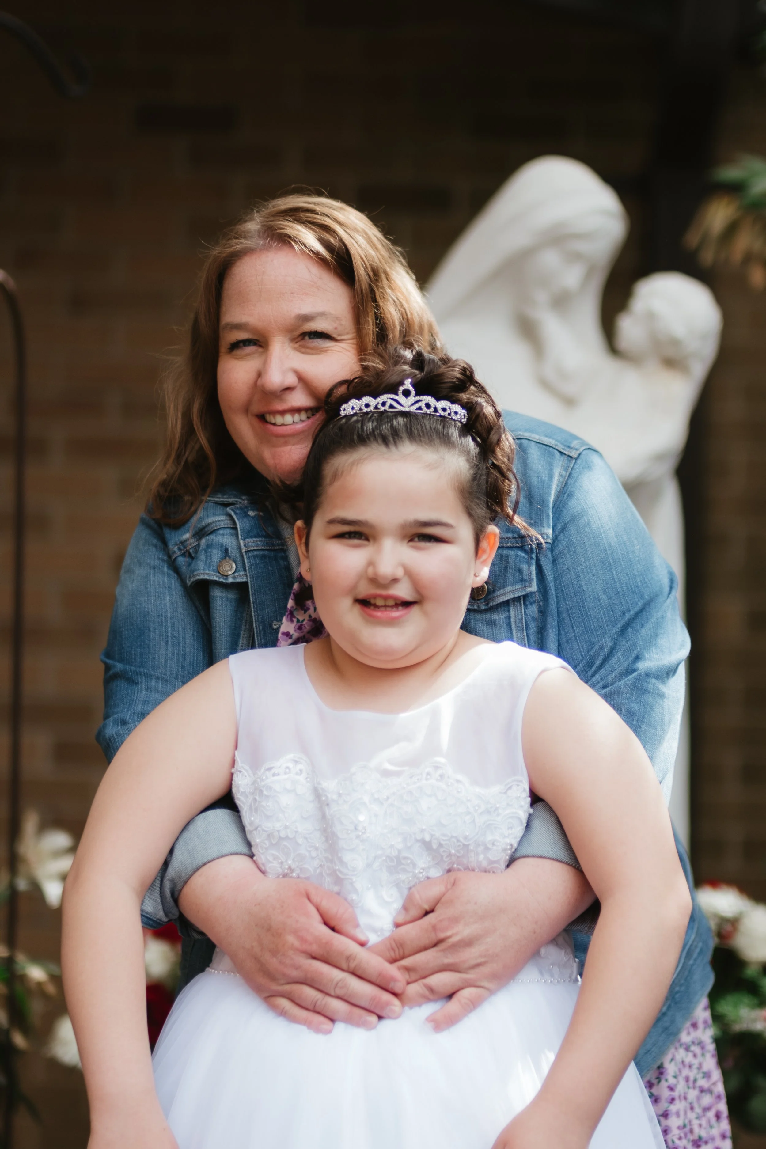 A woman and a young girl smiling, with the woman embracing the girl from behind. The girl is wearing a white dress and a tiara. The background features a sculpture of a woman holding a child, and some flowers.
