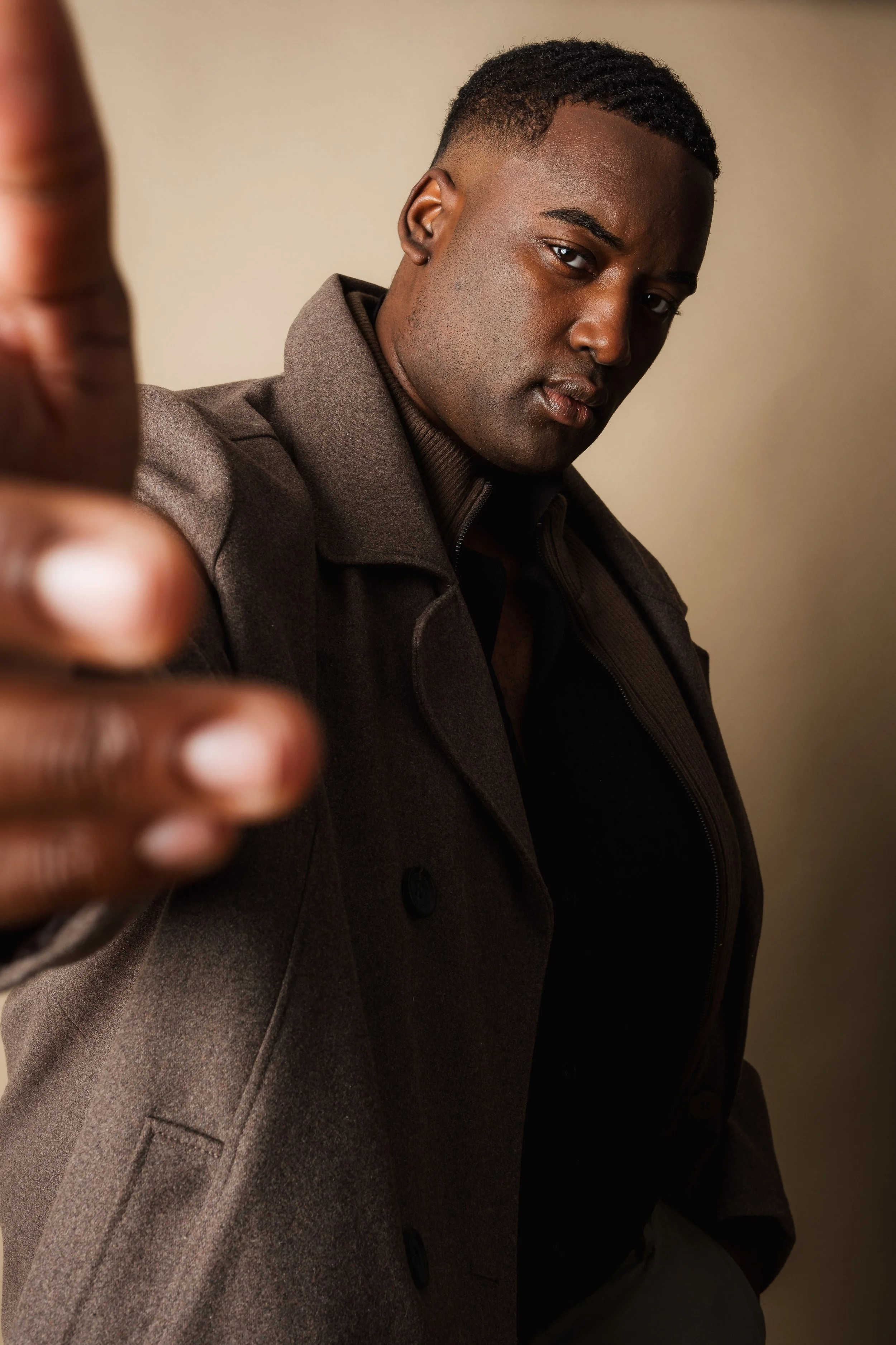 A young man with dark skin and short hair is wearing a brown coat and black shirt, pointing towards the camera with a serious expression.