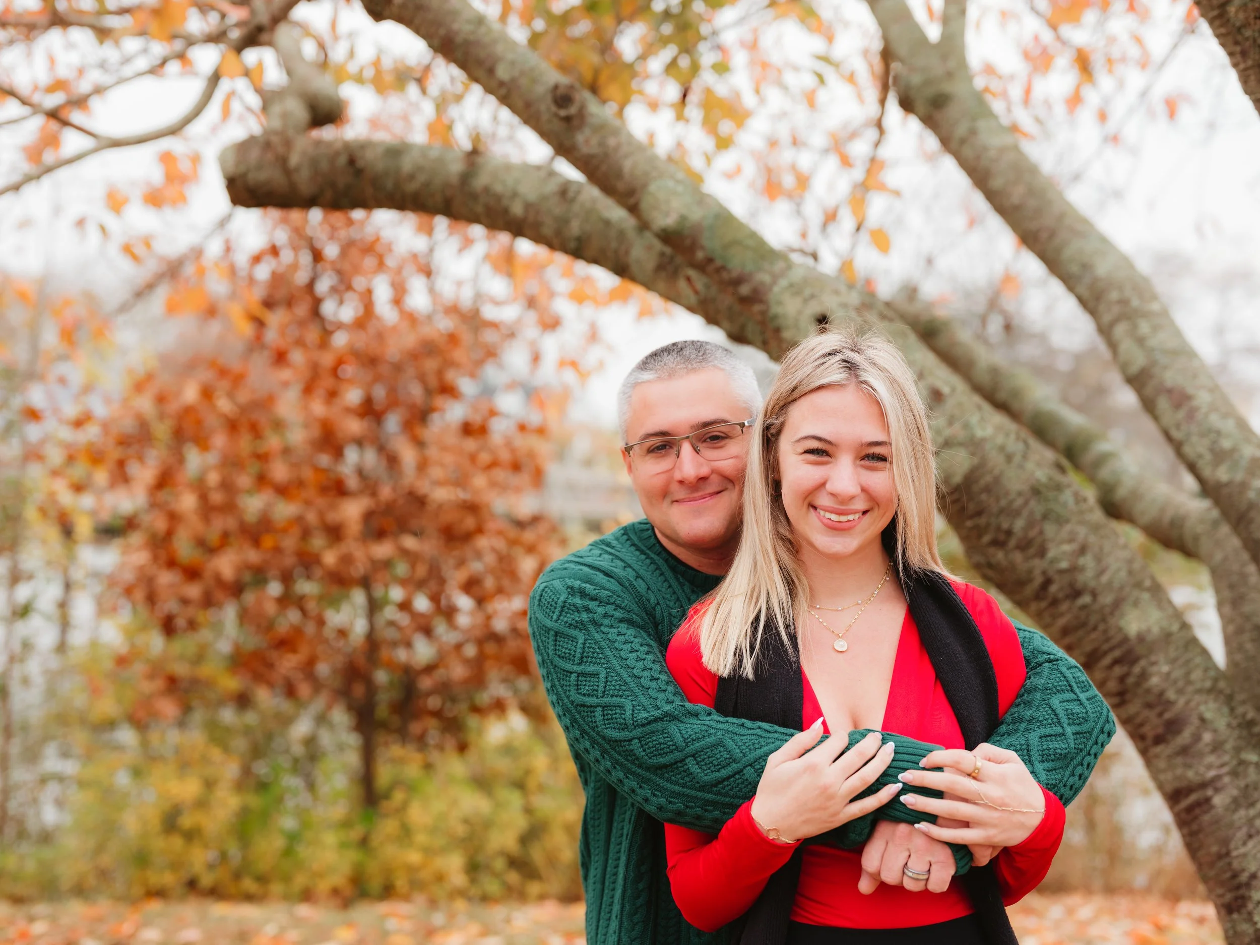 A smiling couple hugging outdoors among autumn trees with orange leaves.