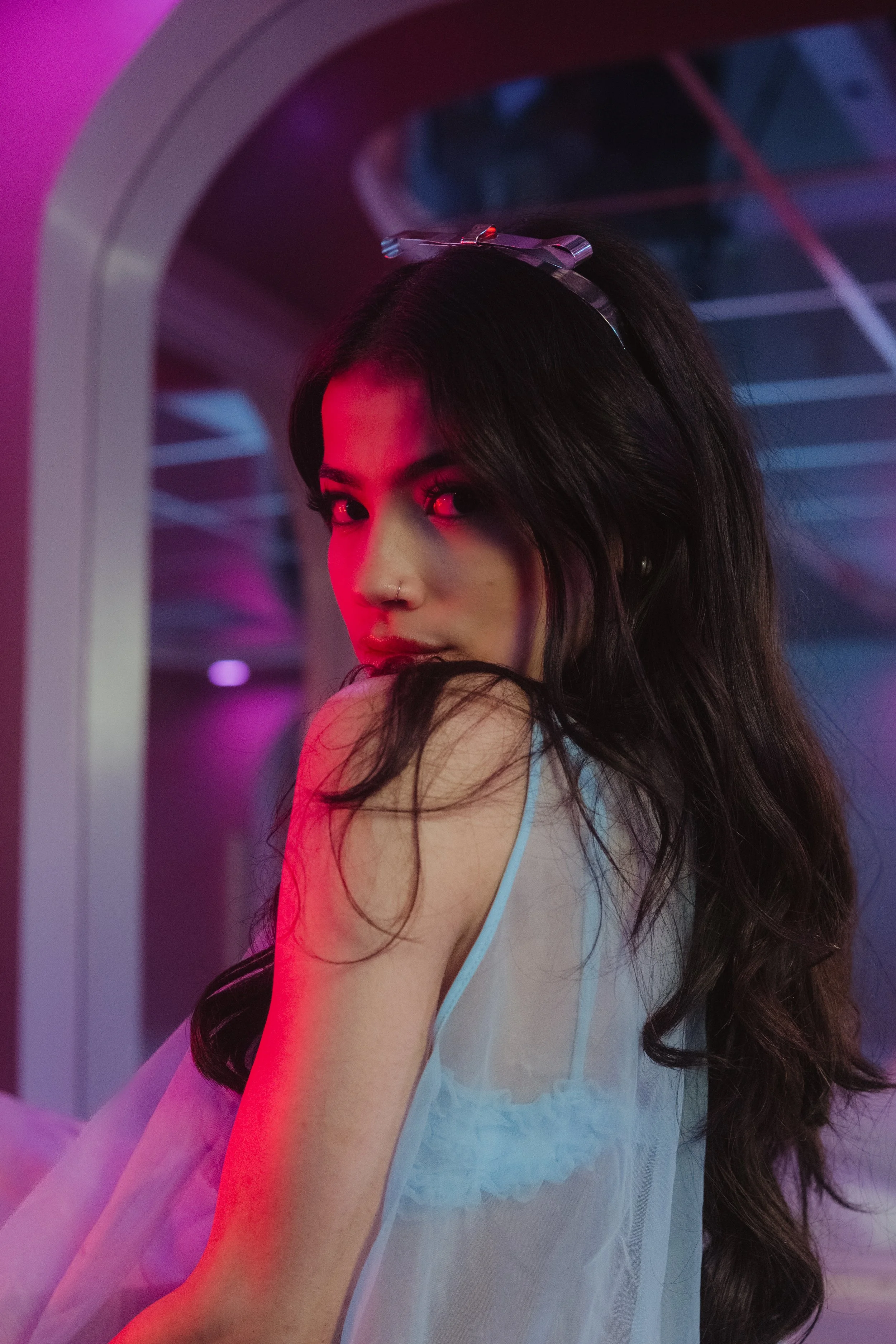A woman with long dark hair and a nose piercing looks over her shoulder in a room with pink and purple lighting.