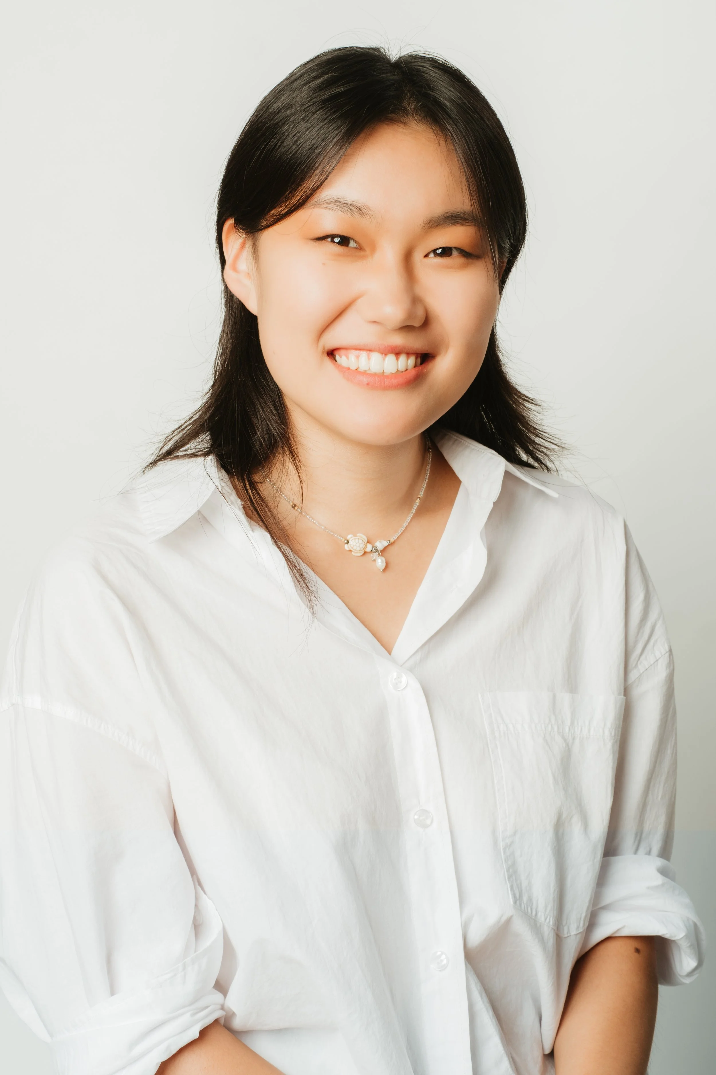 A young woman with shoulder-length black hair smiling, wearing a white button-up shirt and a delicate necklace, against a plain white background.