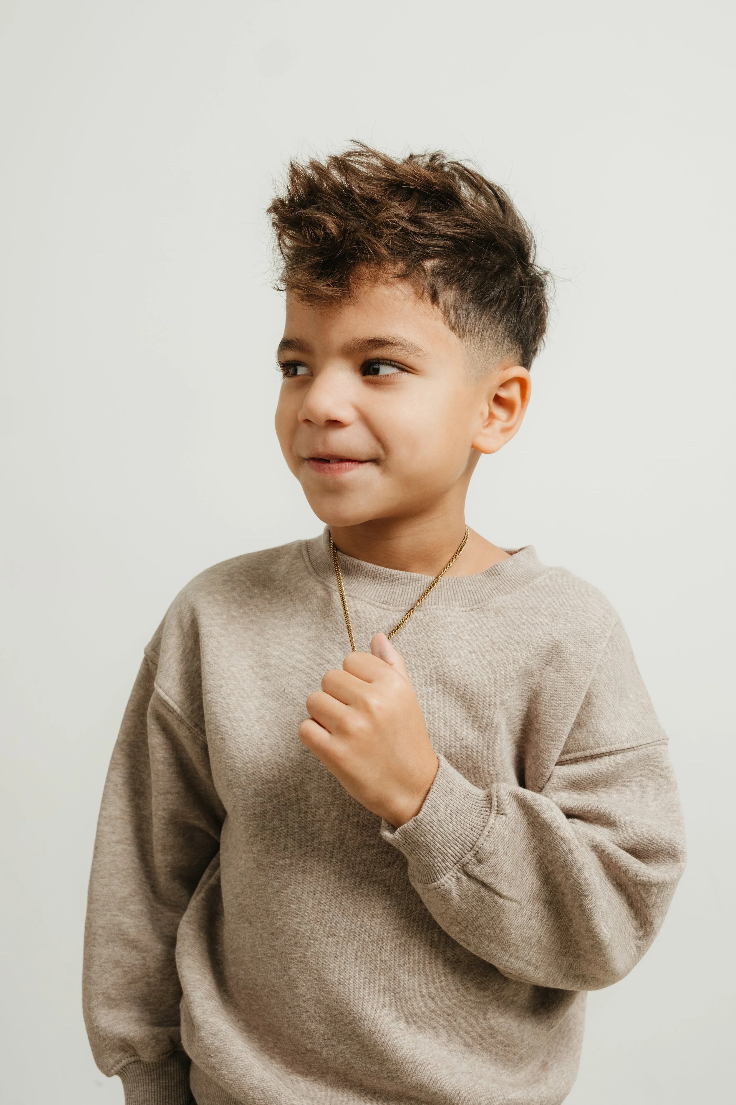 A young boy with short, curly hair and light skin, wearing a beige sweatshirt and a gold chain necklace, standing against a plain white wall.