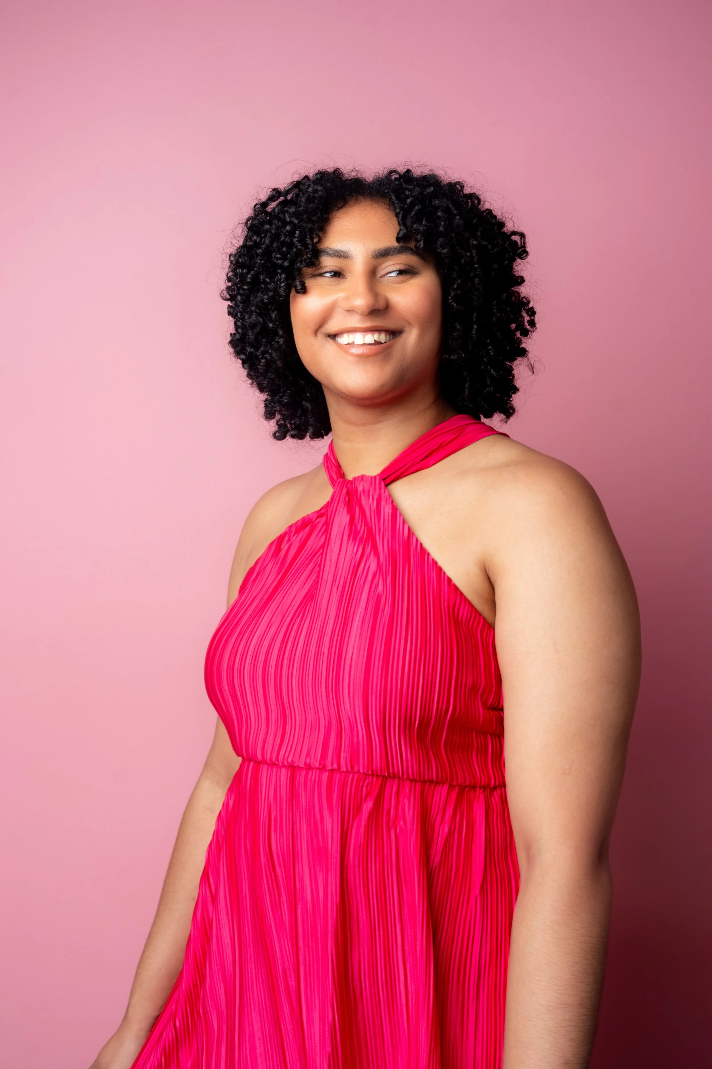 A woman with curly black hair wearing a bright pink, sleeveless, pleated dress standing against a pink background, smiling and looking slightly to her left.
