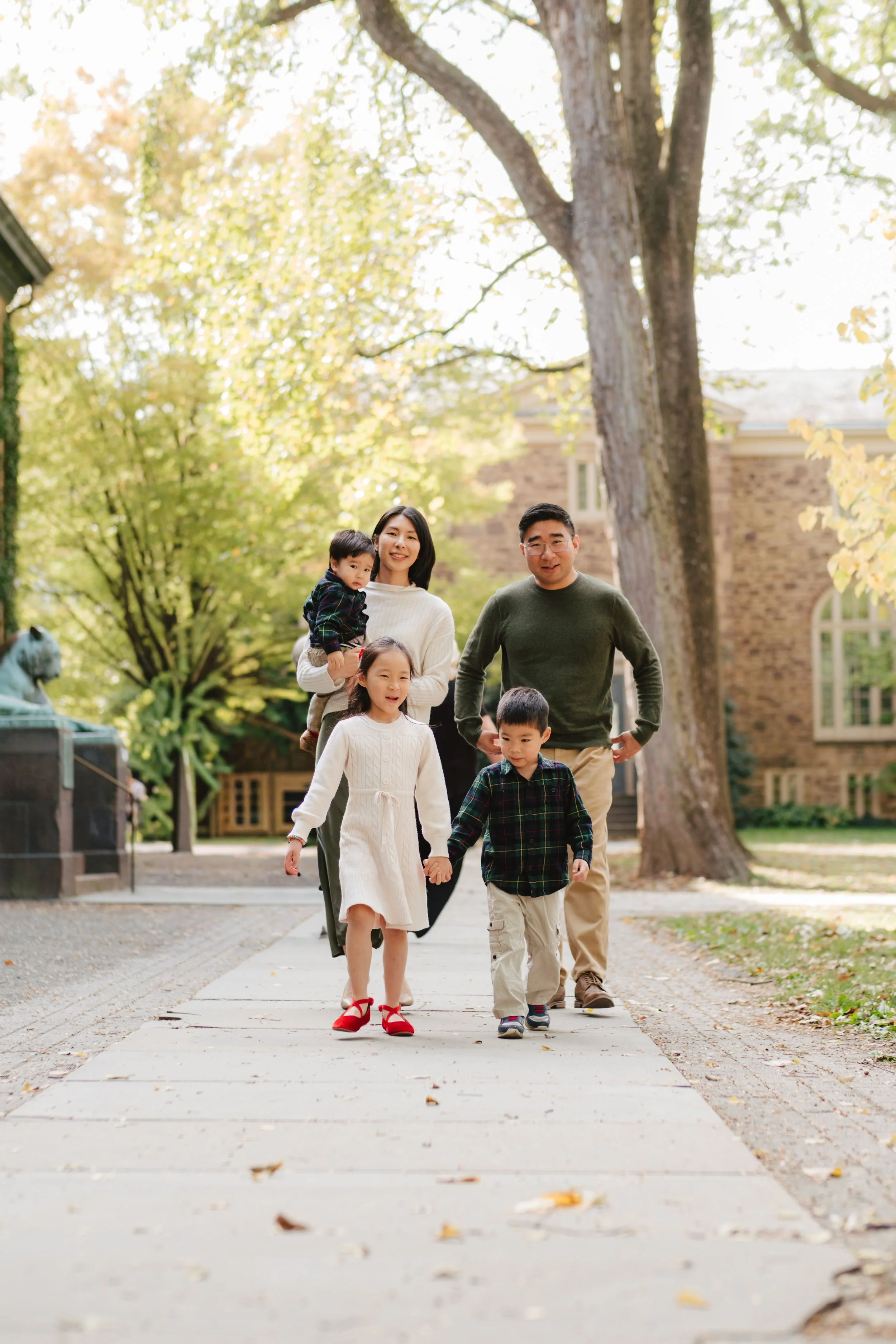 A family of five, including two children, walking together on a sidewalk in a park with trees and a brick building in the background.