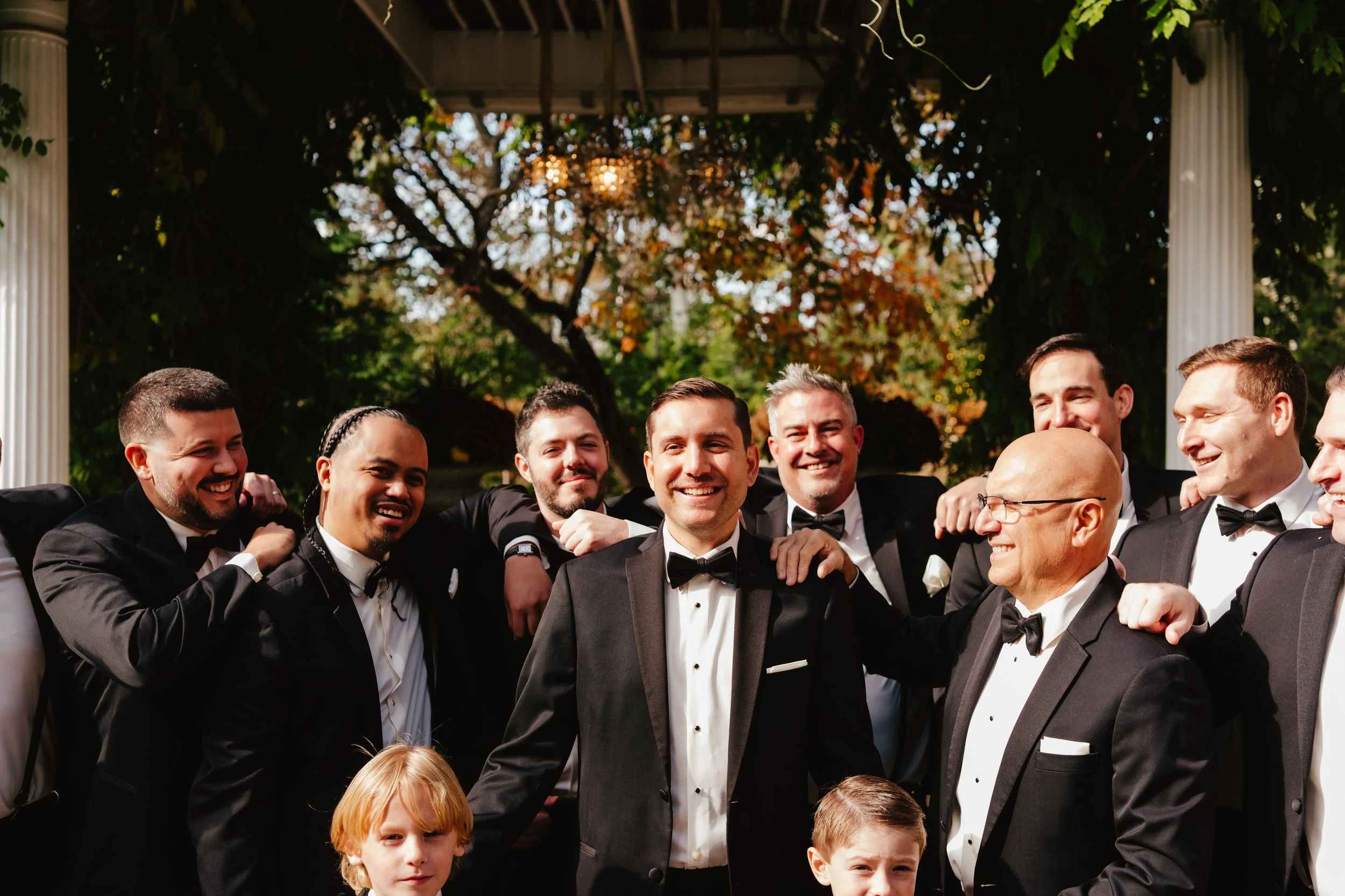 Group of men and two children in formal tuxedos and dresses, smiling and posing outdoors with trees and a structure in the background.
