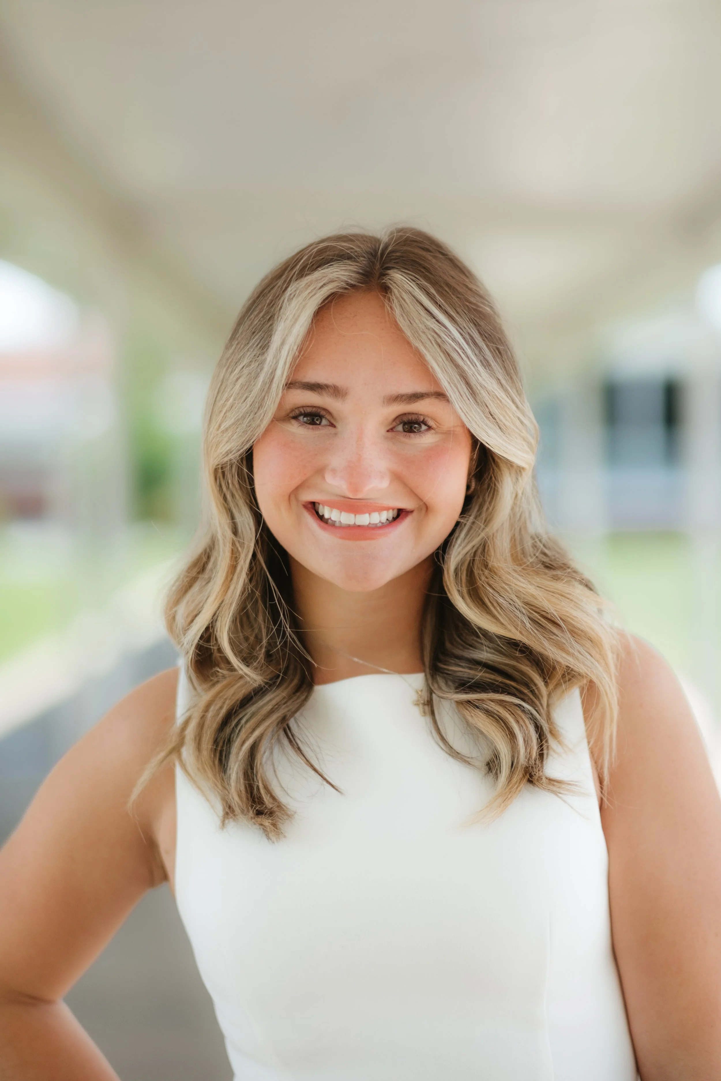 Portrait of a young woman with blonde wavy hair, smiling, wearing a sleeveless white top, outdoors with blurred greenery and building in background.