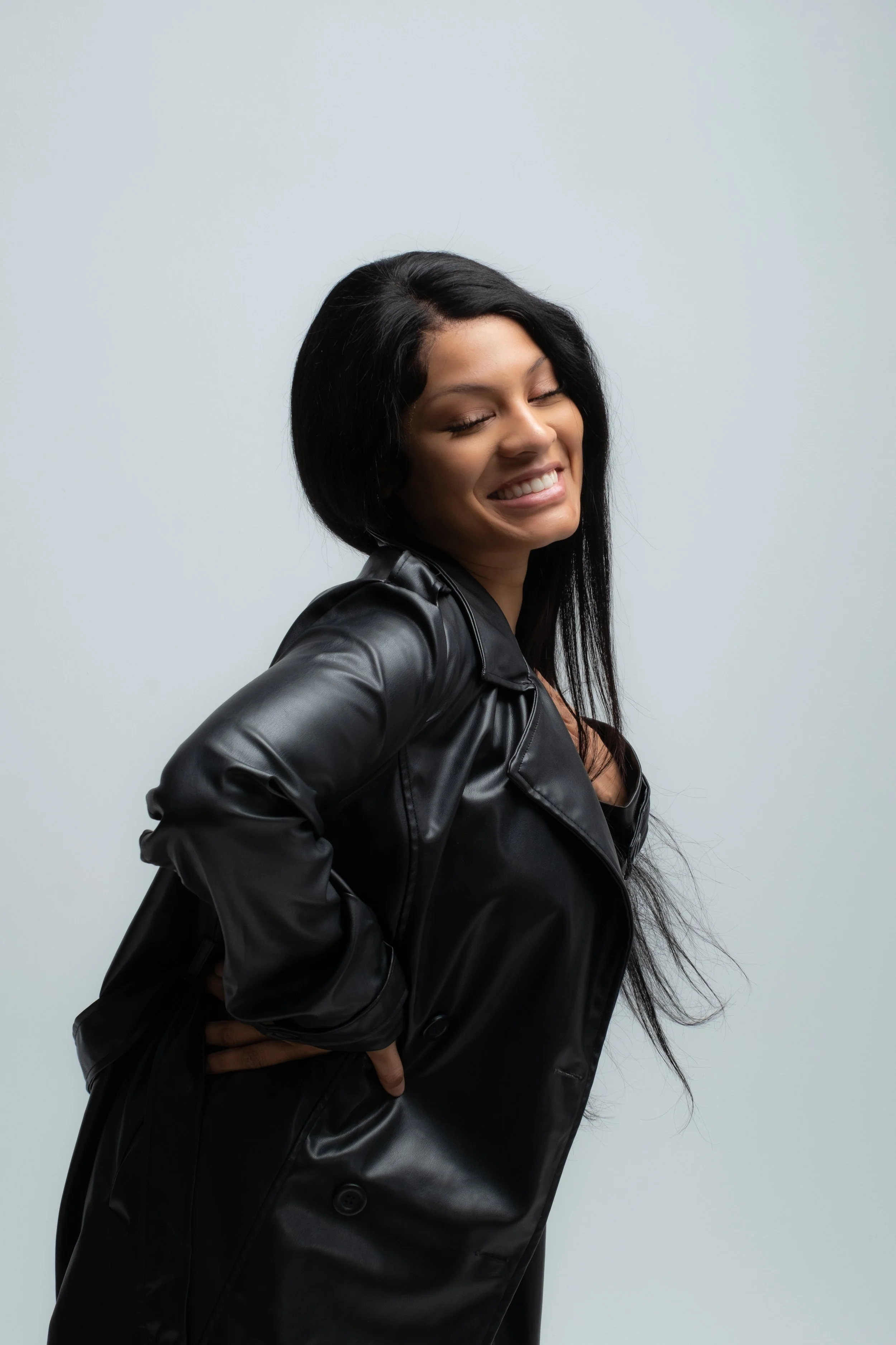 Smiling woman with closed eyes wearing a black leather jacket against a plain light-colored background.