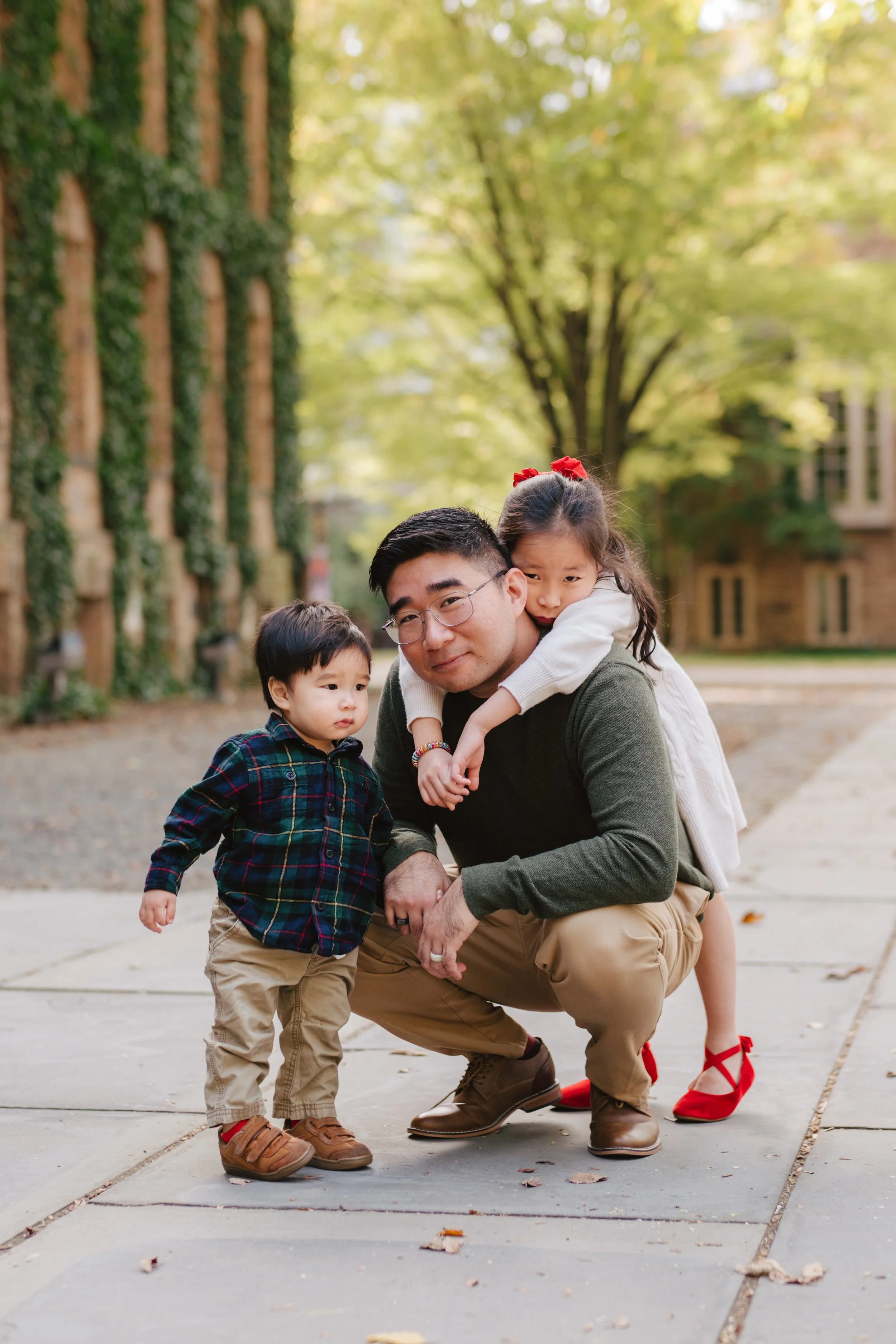 A man crouches on a sidewalk with a young girl on his back and a toddler boy standing beside him outdoors in a park with green trees and buildings in the background.