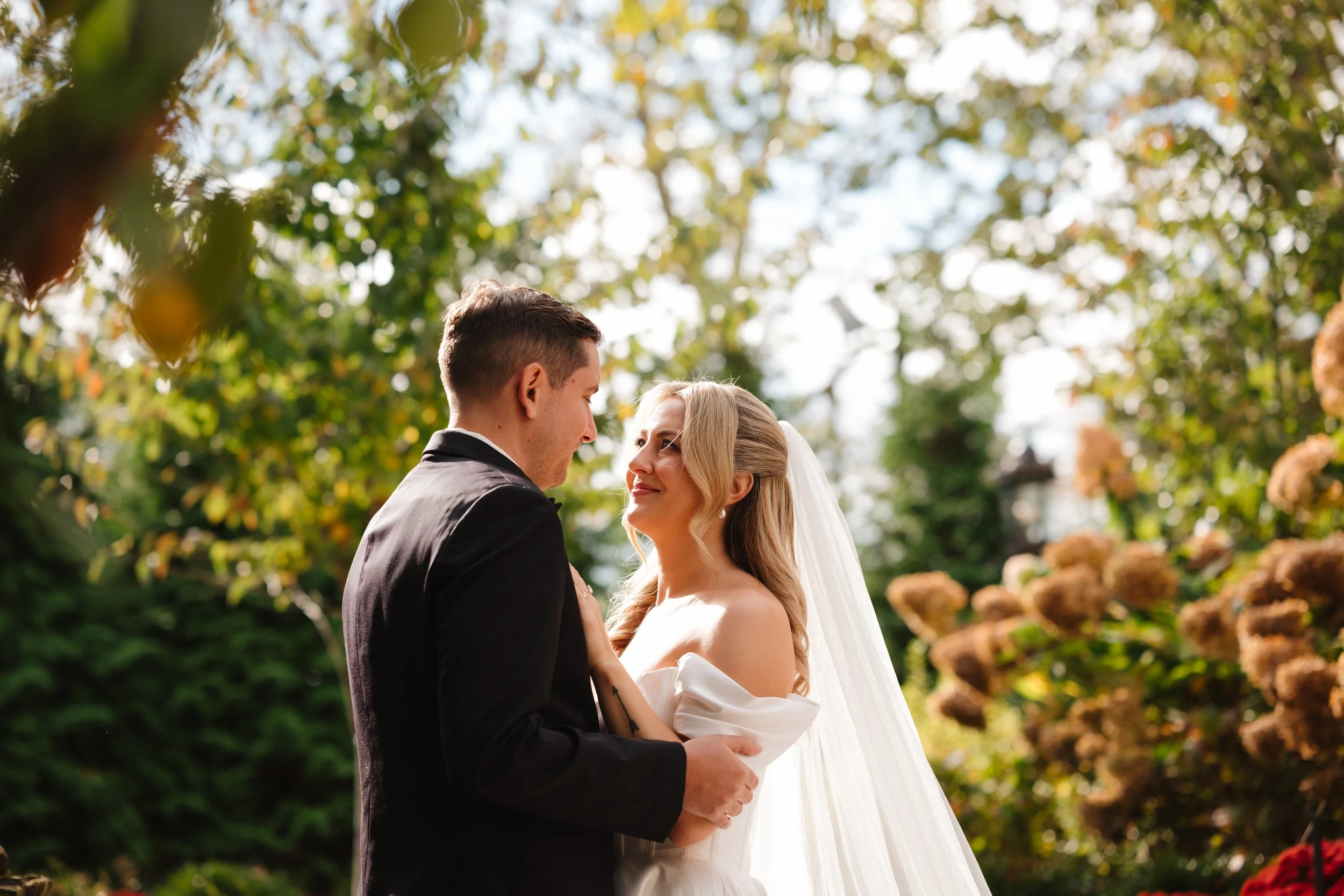 Bride and groom sharing a romantic moment outdoors during their wedding, surrounded by greenery and flowers, with sunlight filtering through the trees.