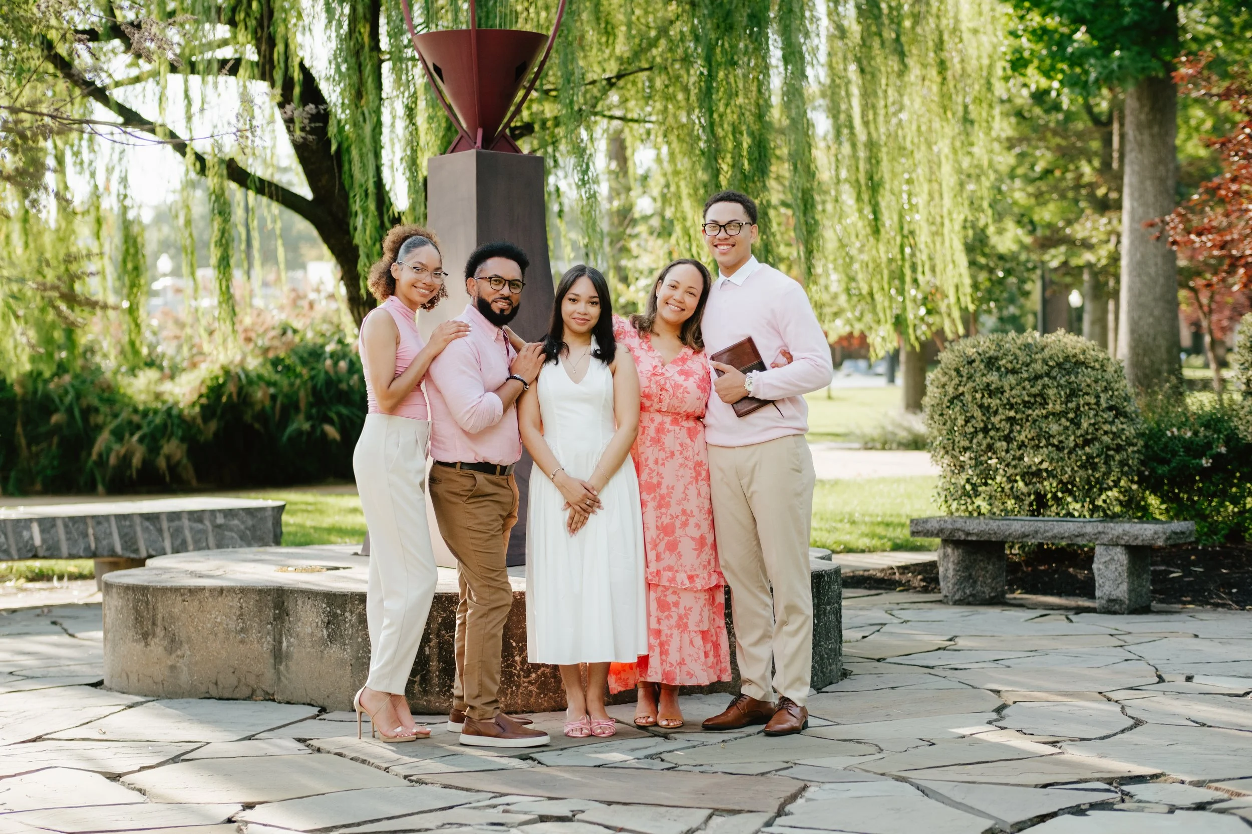 A diverse group of five young adults standing outdoors on a stone path in front of a modern sculpture and lush green trees, smiling at the camera.