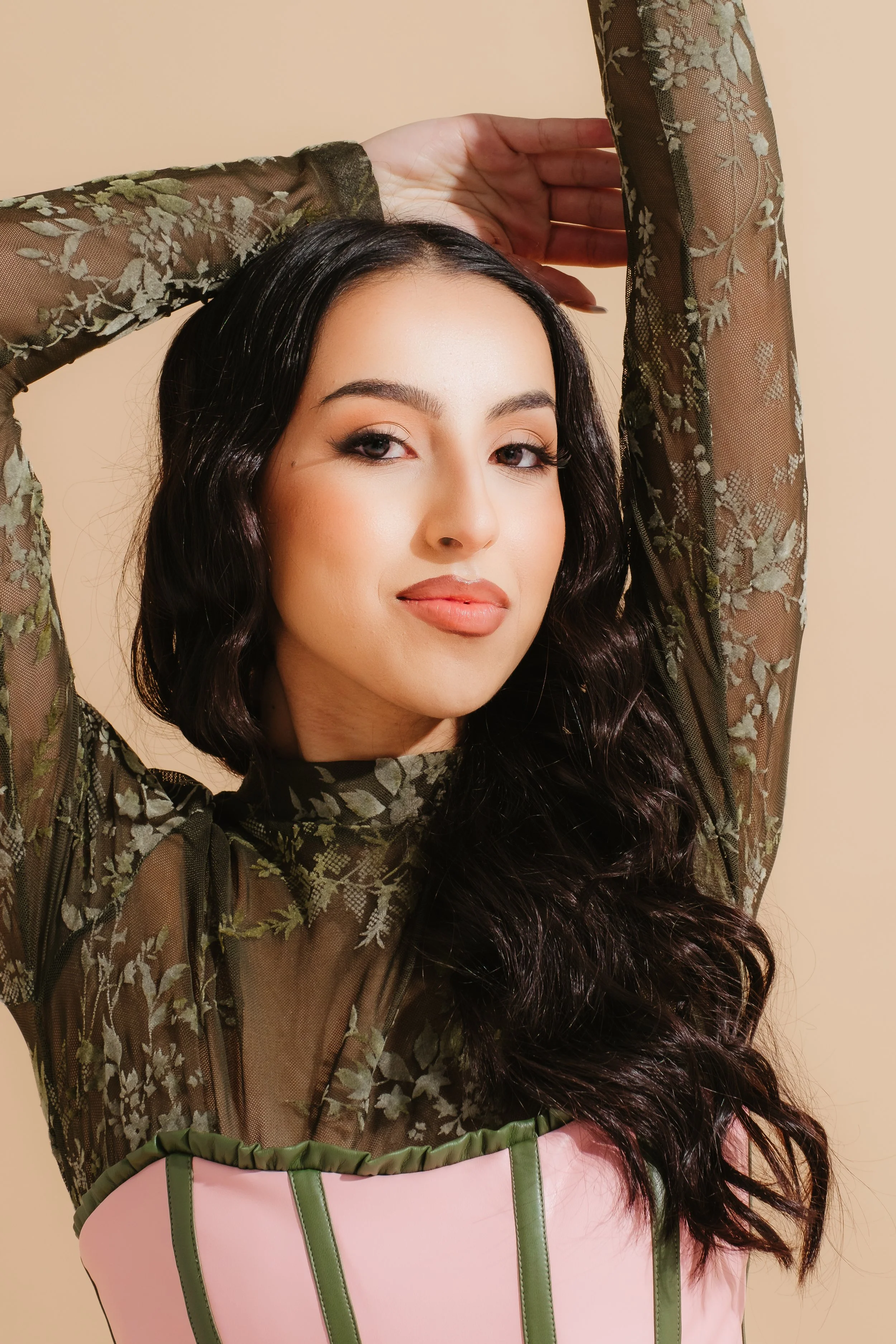 A woman with long, wavy dark hair, fair skin, and makeup, wearing a sheer, green, floral-patterned top and a pink corset with green accents, standing against a plain beige background.