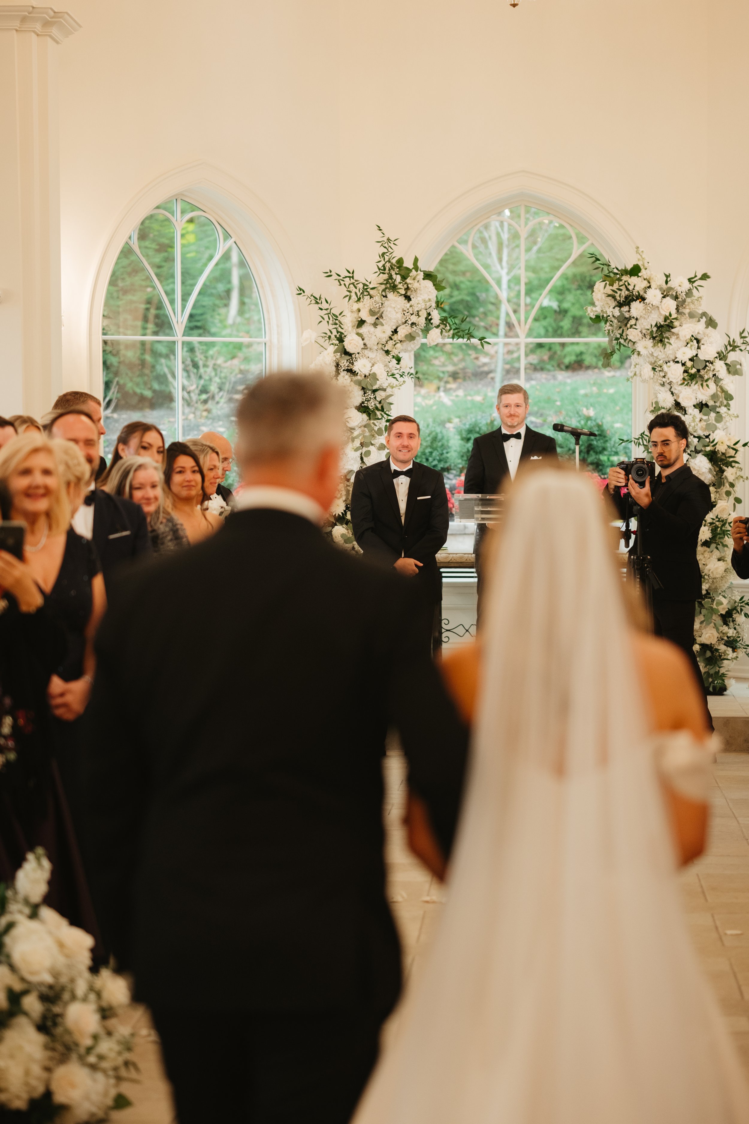 A wedding ceremony inside a church with two grooms standing at the altar, surrounded by guests, floral arrangements, and large arched windows.