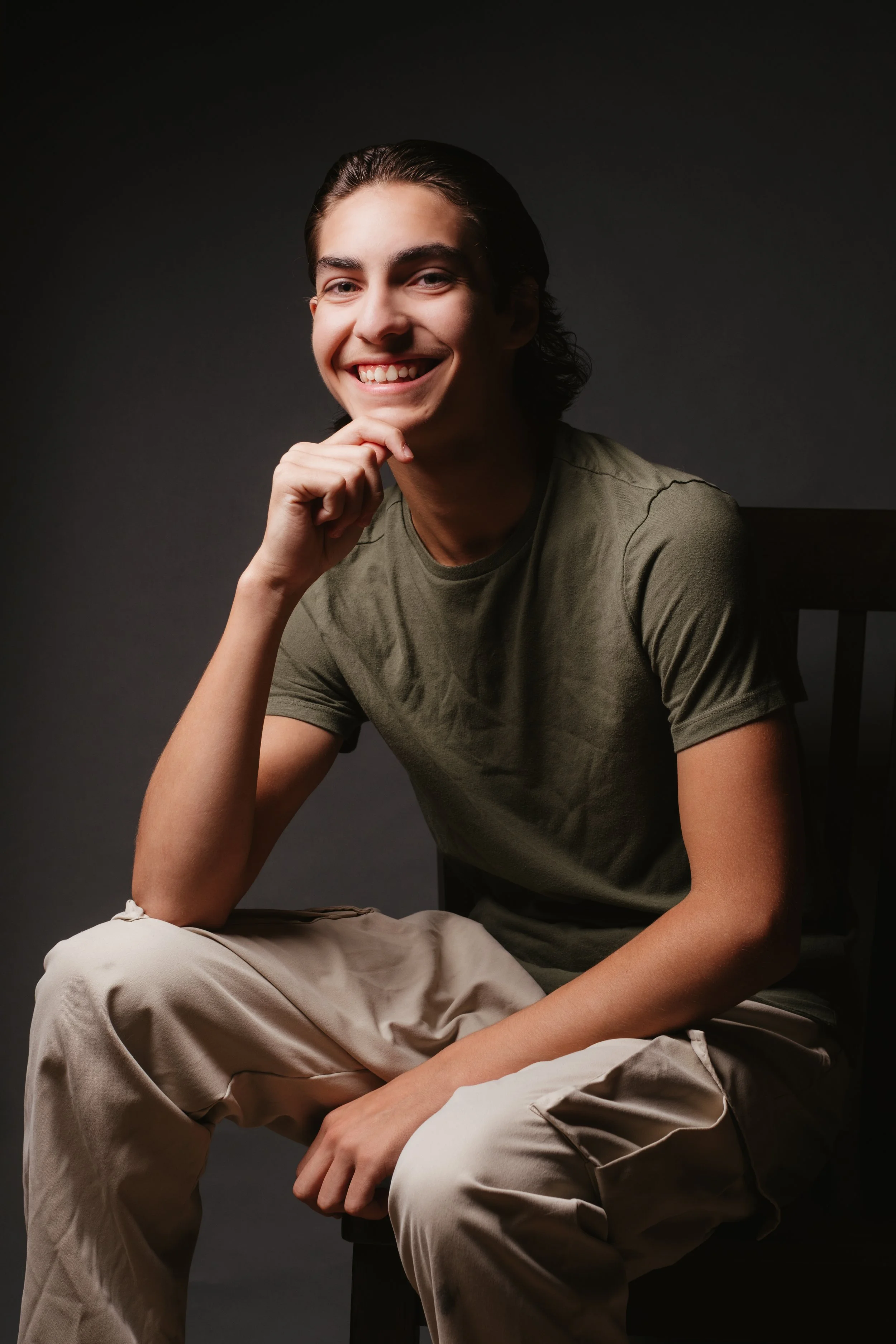 A young man with dark hair, smiling, sitting on a chair against a dark background.
