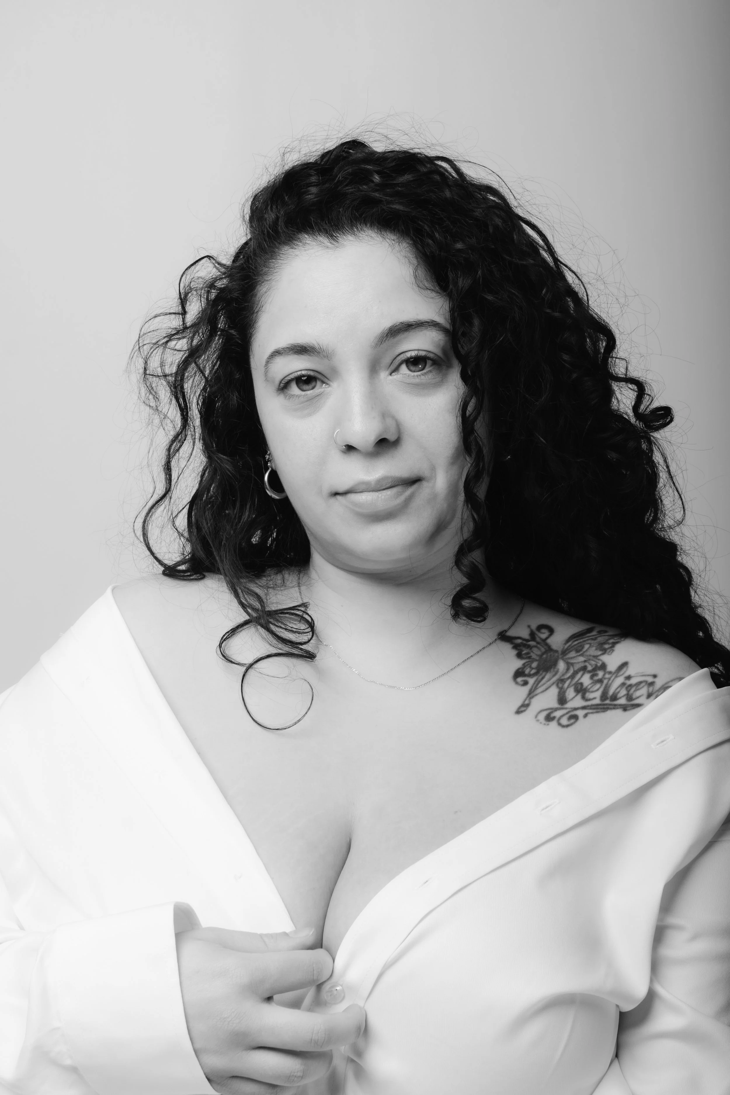 Black and white portrait of a woman with curly hair, wearing a white shirt off the shoulder, holding it with one hand, with a tattoo on her shoulder that says 'Believe' and a butterfly. She has earrings, a nose ring, and a delicate necklace.