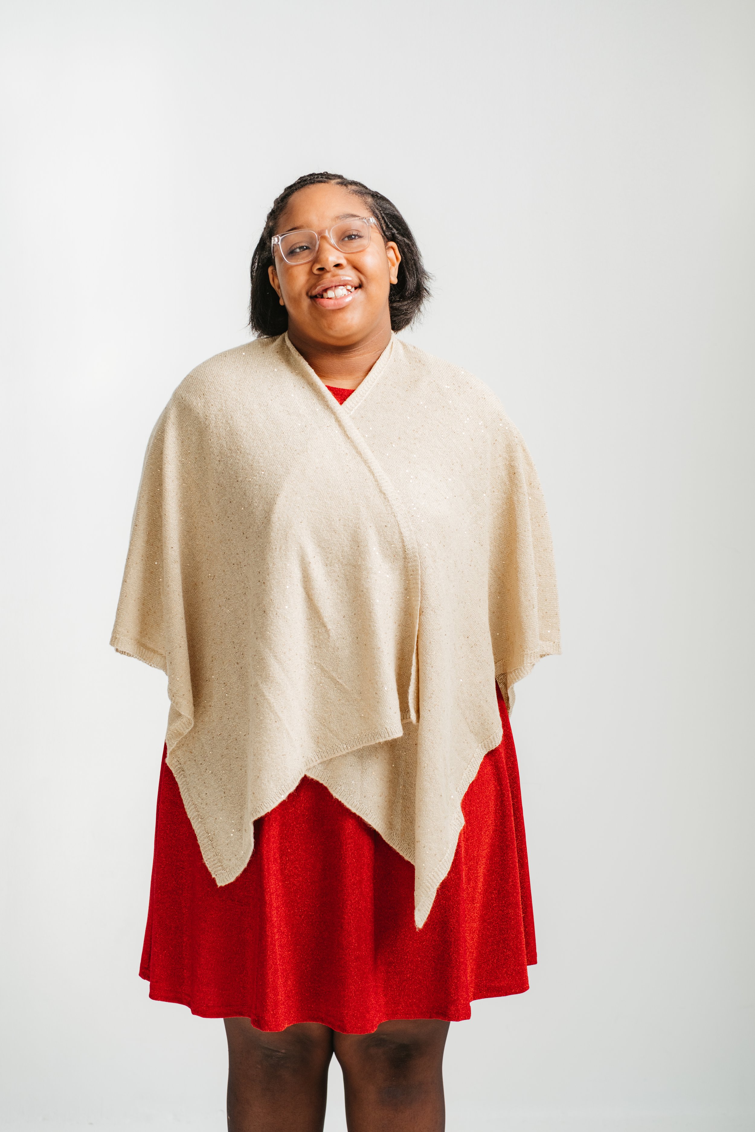 A young girl with glasses, wearing a beige shawl over a red dress, standing against a white background.