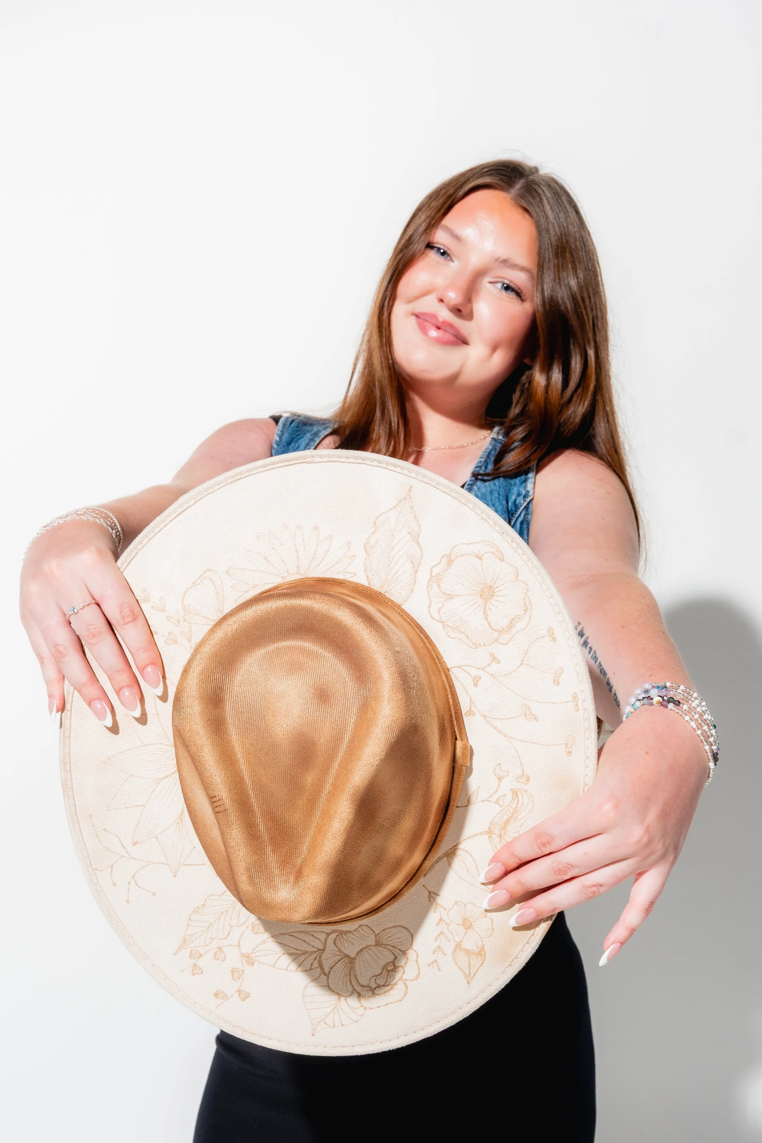 A young woman with long brown hair holding a large beige hat with a gold ribbon and floral embroidery