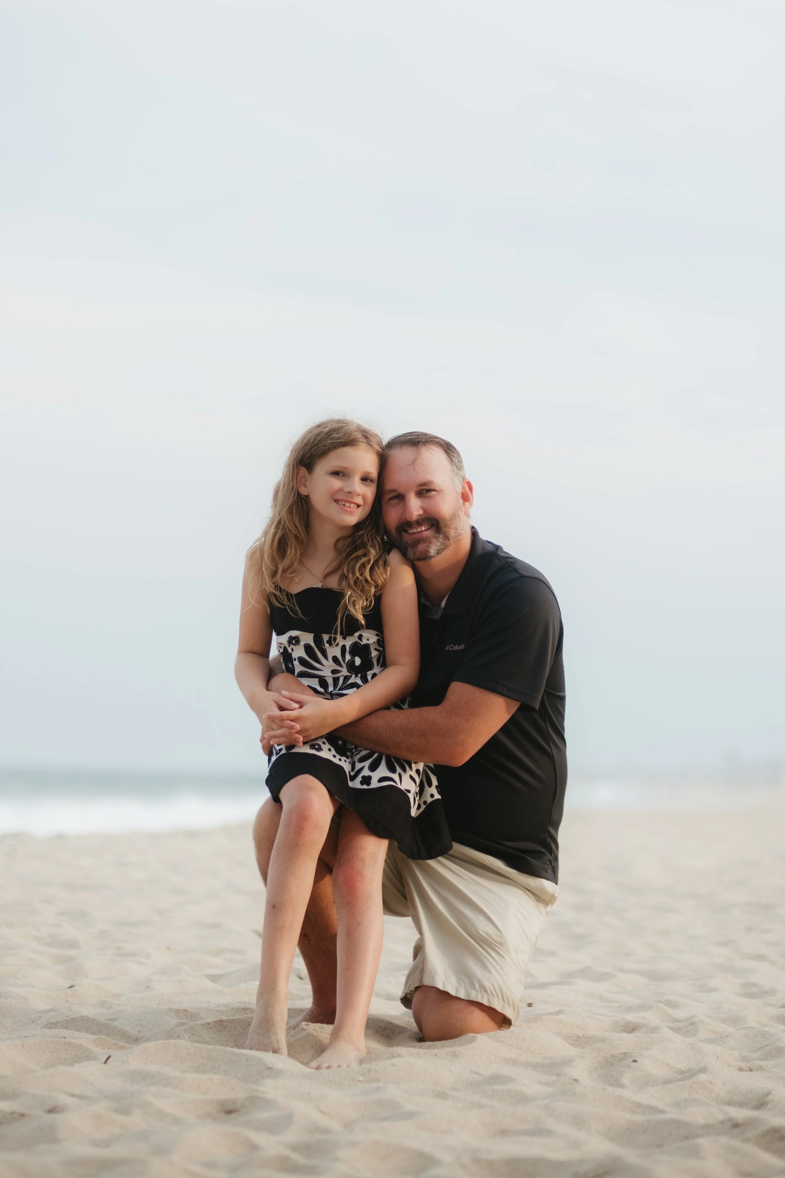 A man and a young girl kneeling on the sand at the beach, smiling for a photo with the ocean in the background.