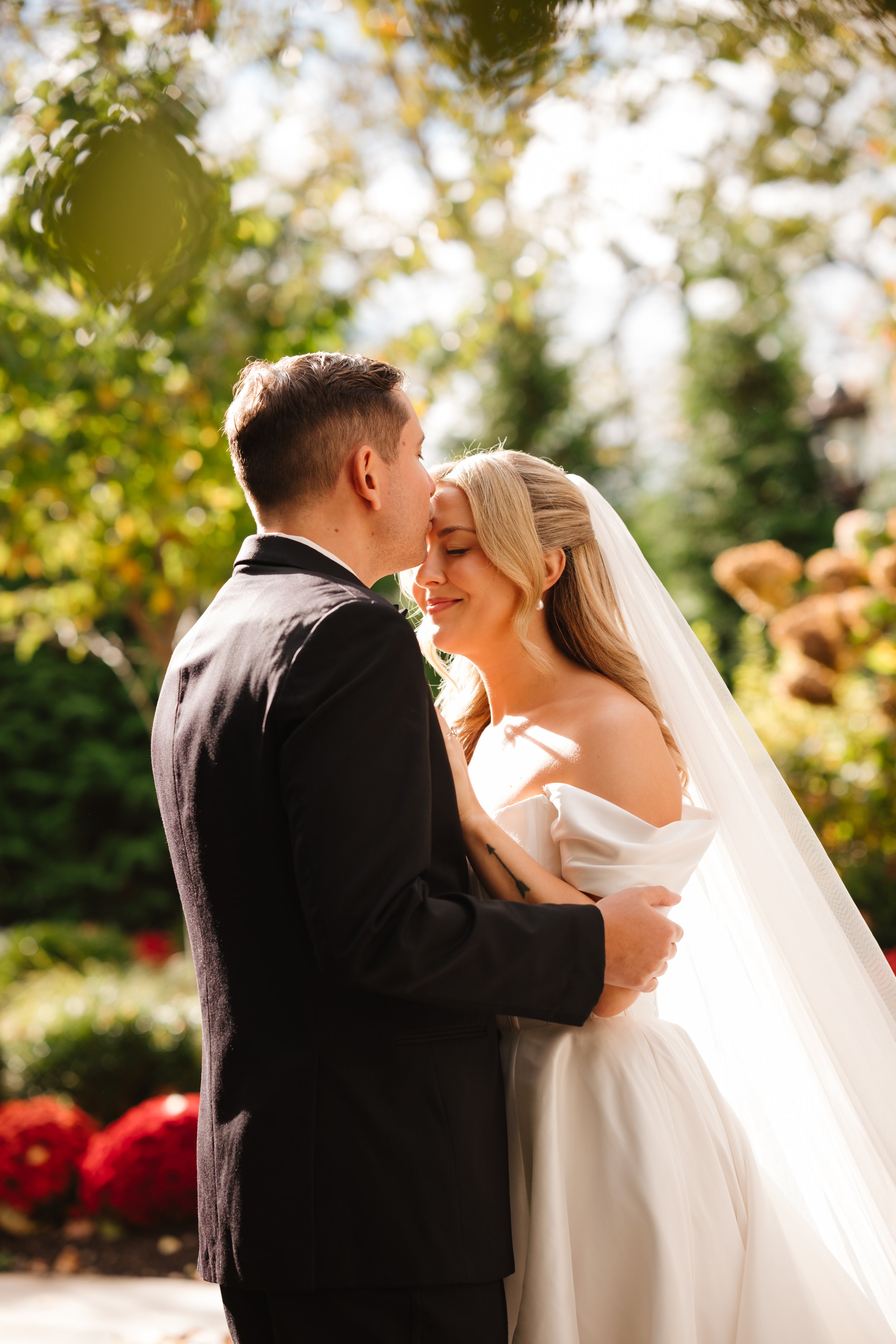A newlywed couple sharing an intimate moment outdoors, with the groom gently kissing the bride on her forehead. The bride is wearing a white wedding dress and veil, and the groom is dressed in a black tuxedo. The background consists of greenery and a