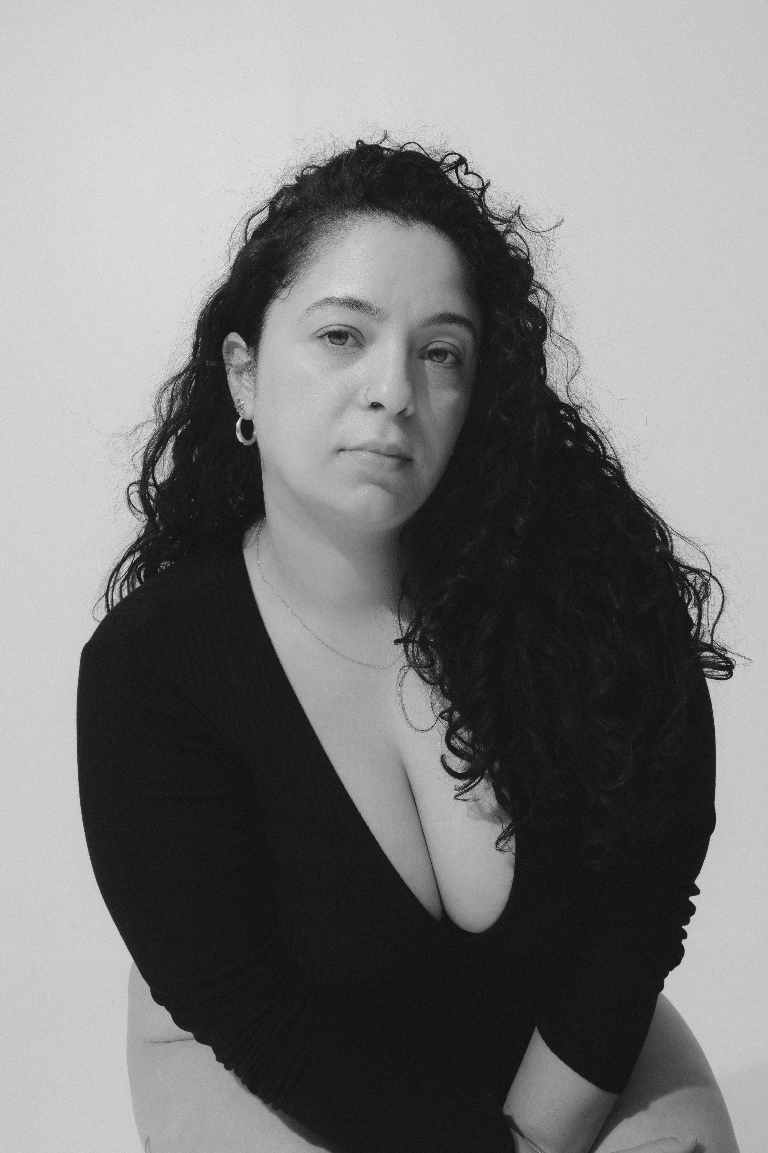 Black and white portrait of a woman with long curly hair, wearing a black top and jewelry, sitting against a plain background.
