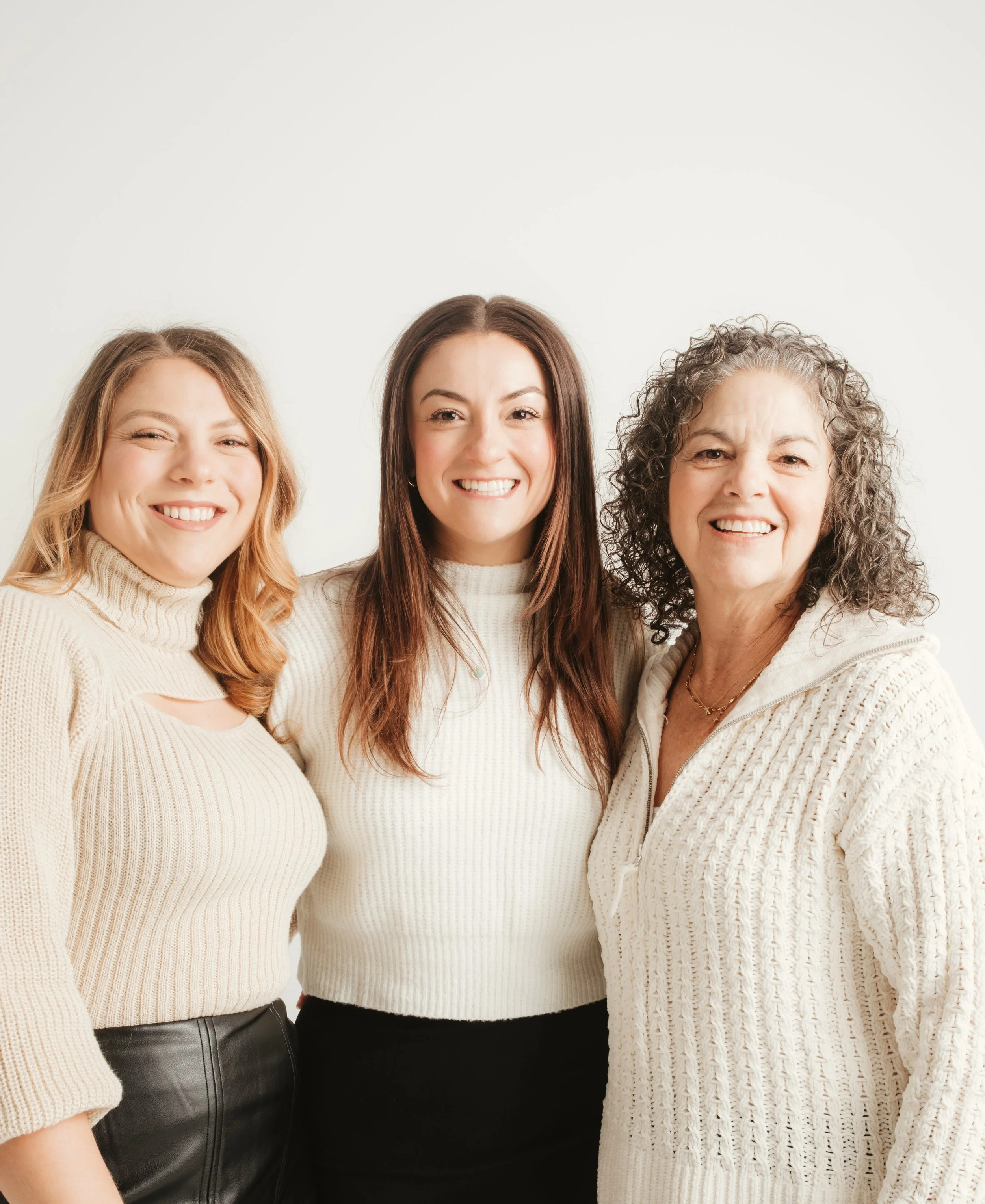 Three women standing close together, smiling, dressed in cozy sweaters, with a plain light background.