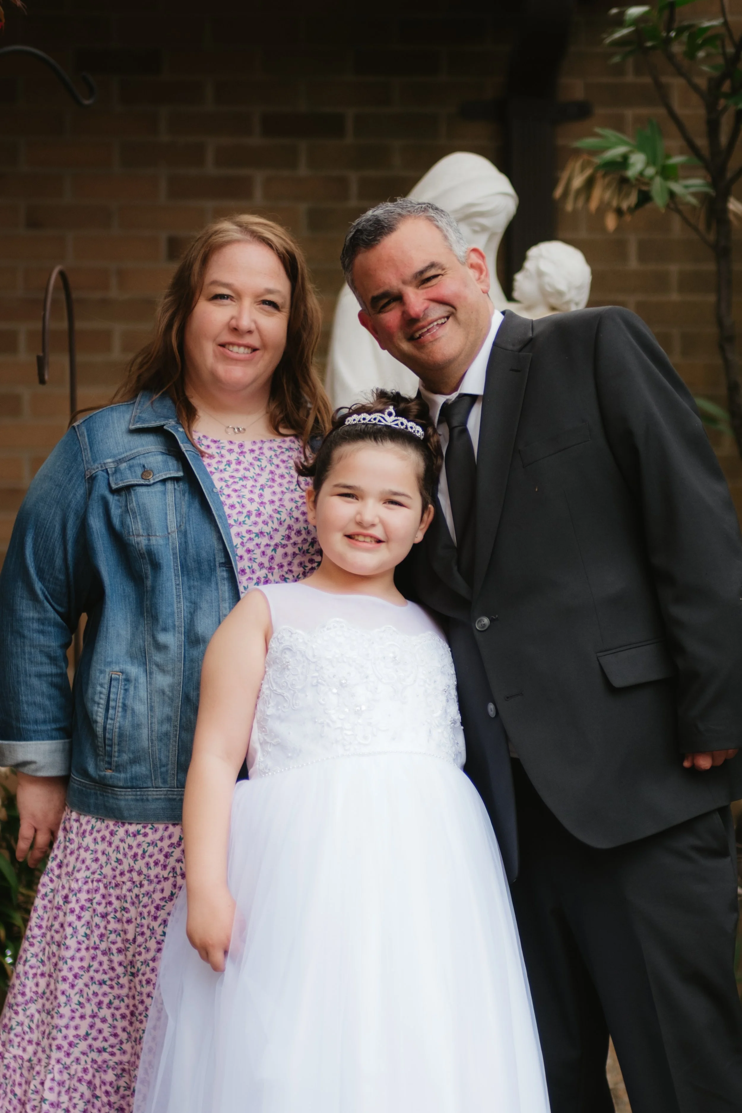 A family photo of three people: a woman, a man, and a girl in a white dress, standing outdoors in front of a brick wall with sculptures and plants.