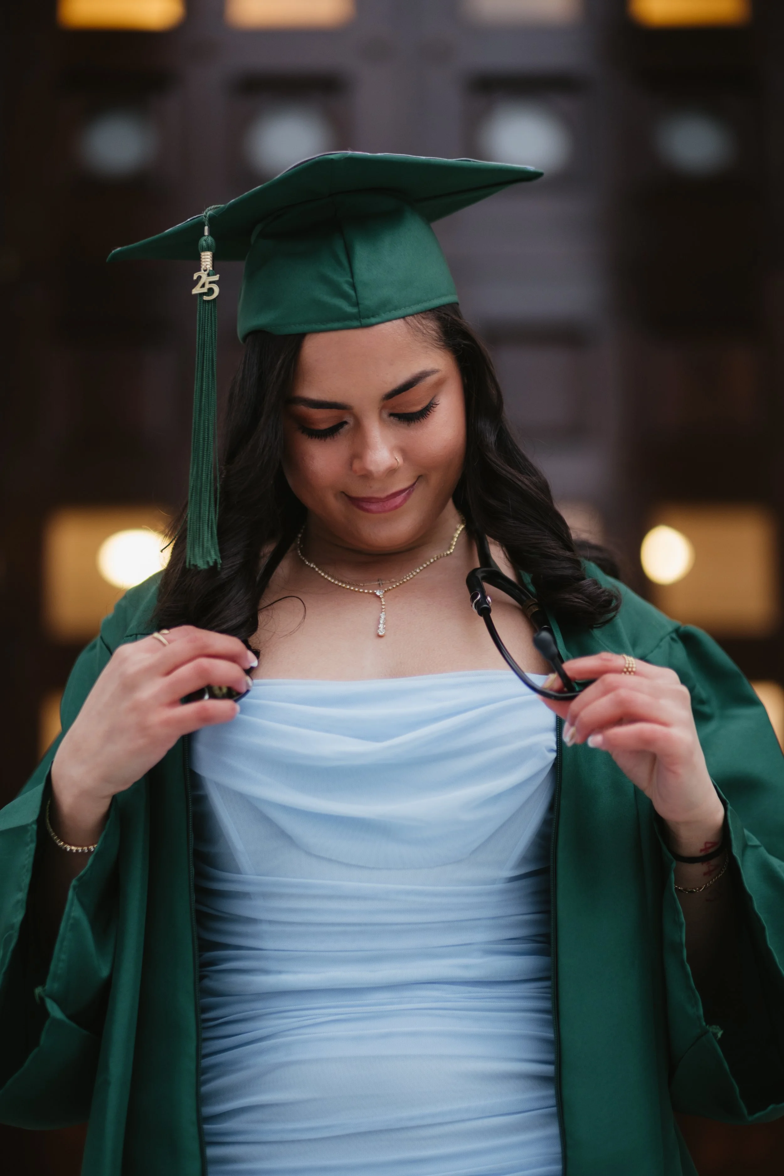 A young woman dressed in a green graduation cap and gown, white dress, holding a stethoscope, standing indoors with blurred lights in the background.