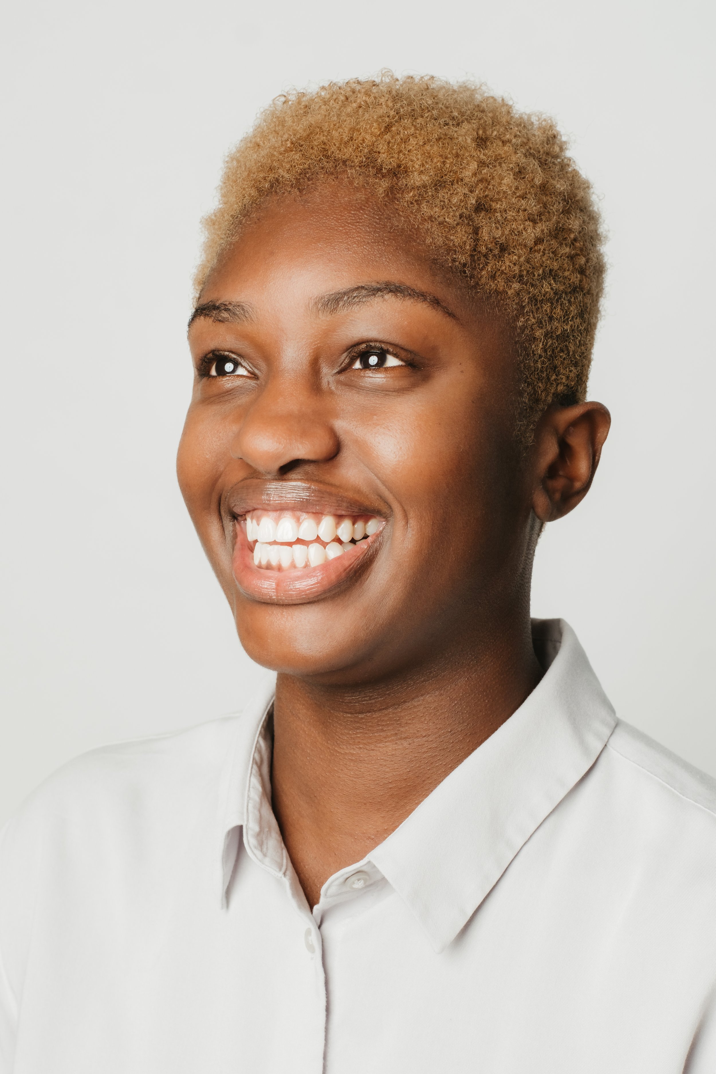 Close-up of a smiling woman with short blonde hair, wearing a white collared shirt, looking slightly to her right against a plain white background.