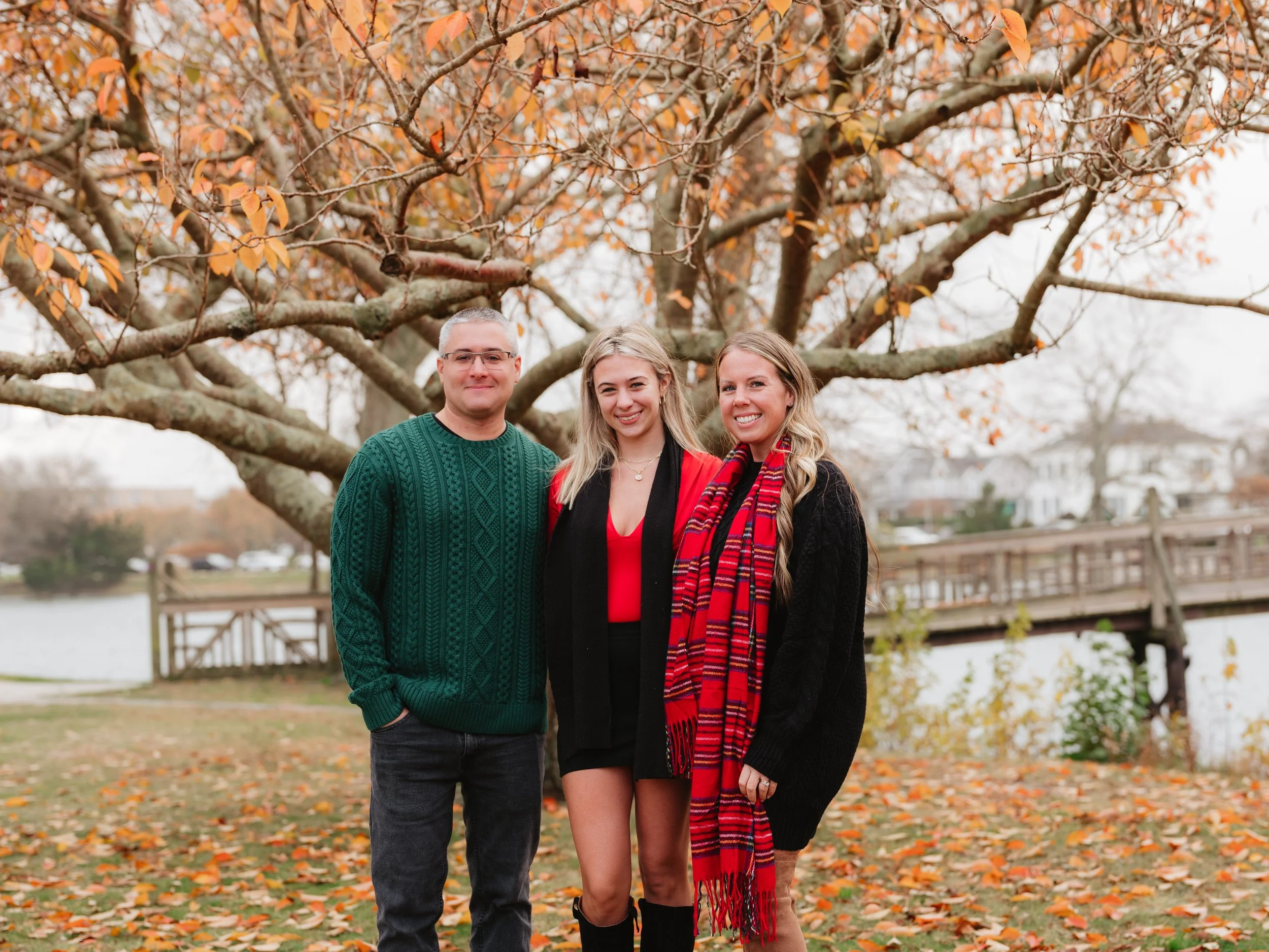Three smiling people standing outdoors on autumn day, in front of a large tree with orange leaves, near a body of water and a wooden bridge.