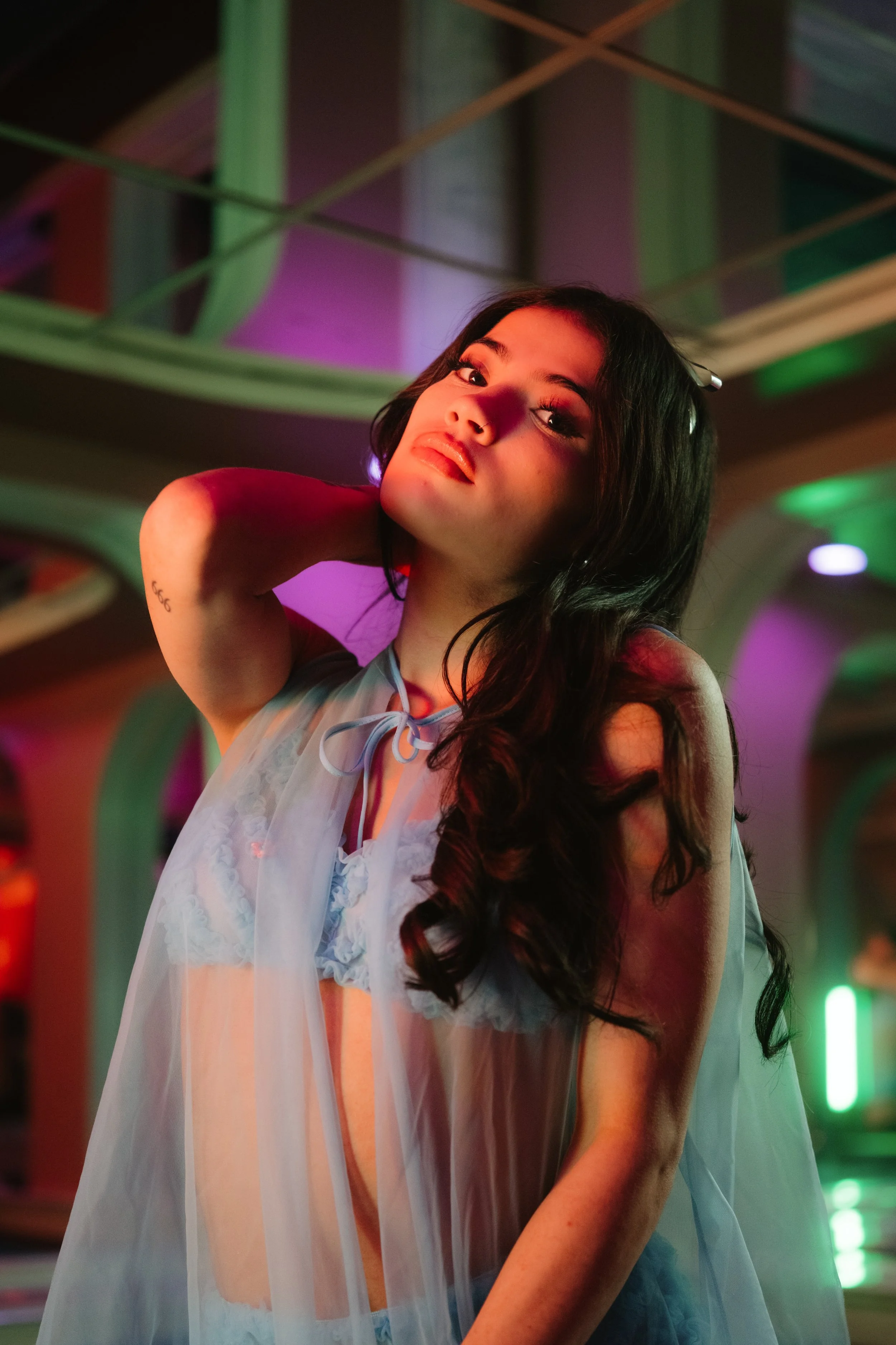 A young woman with long dark hair wearing sheer nightwear, illuminated by colorful neon lights in a vibrant, modern setting.