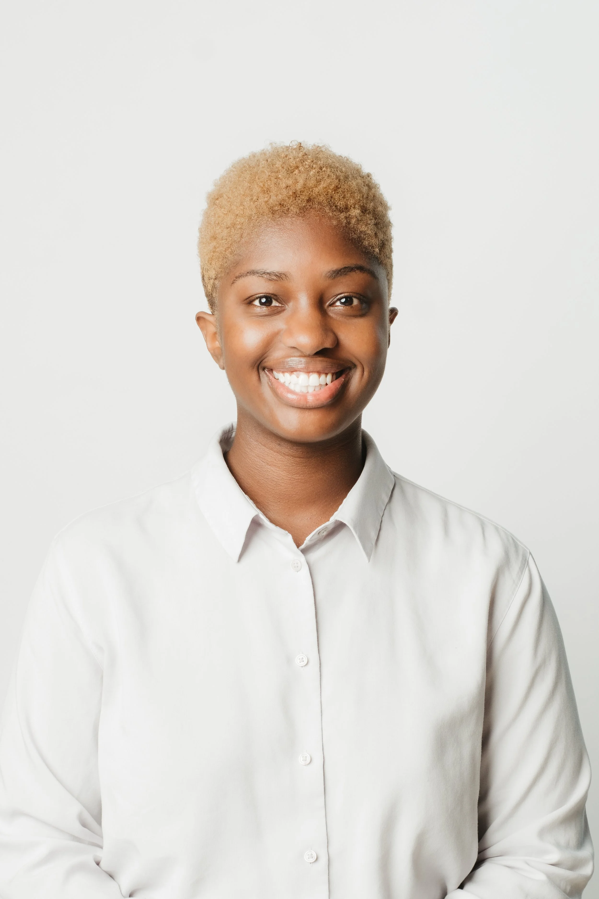 Smiling woman with short curly blonde hair wearing a white shirt against a plain white background.