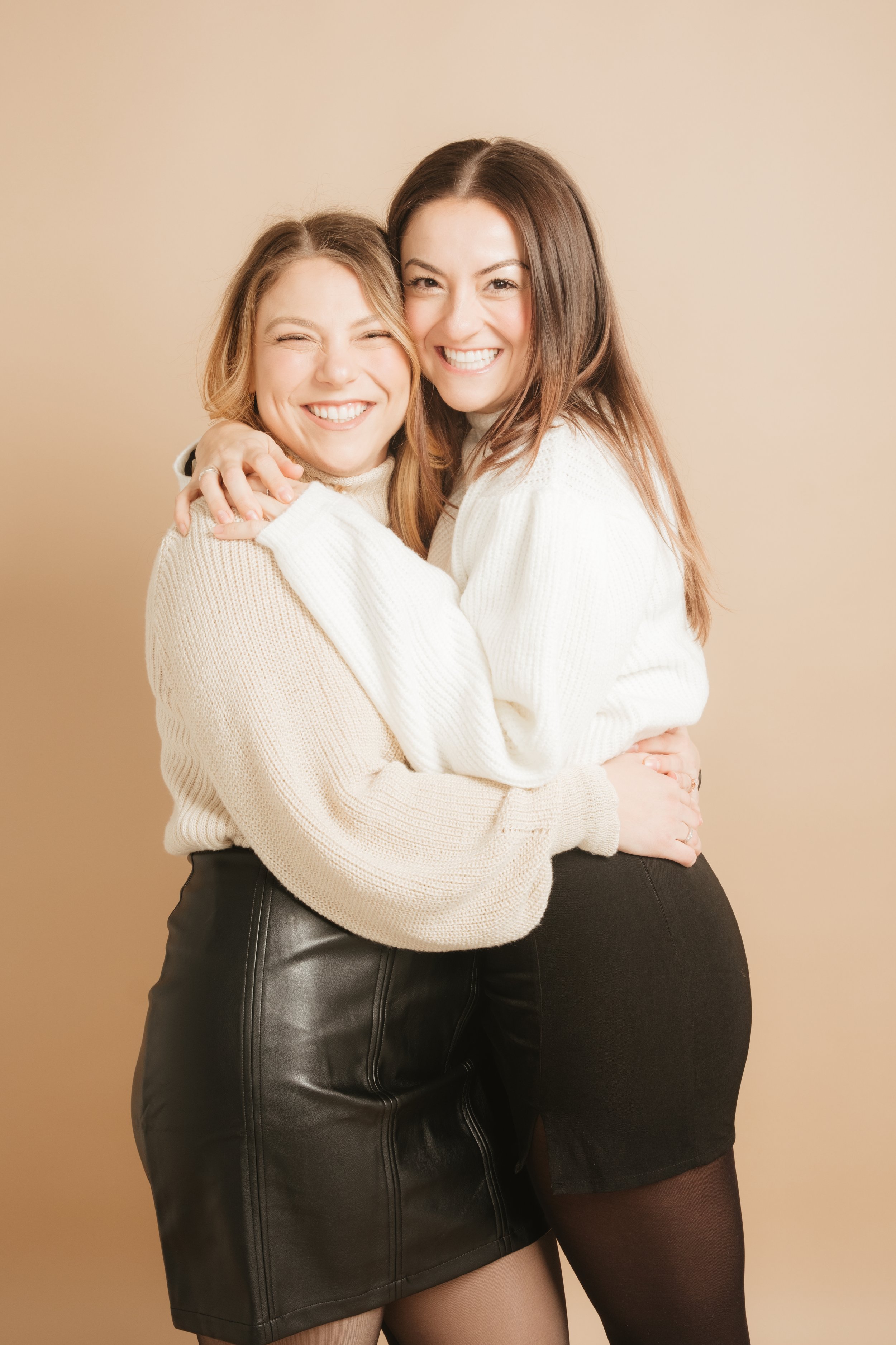 Two women smiling and hugging each other, one with blonde hair and the other with brunette hair, against a neutral background.