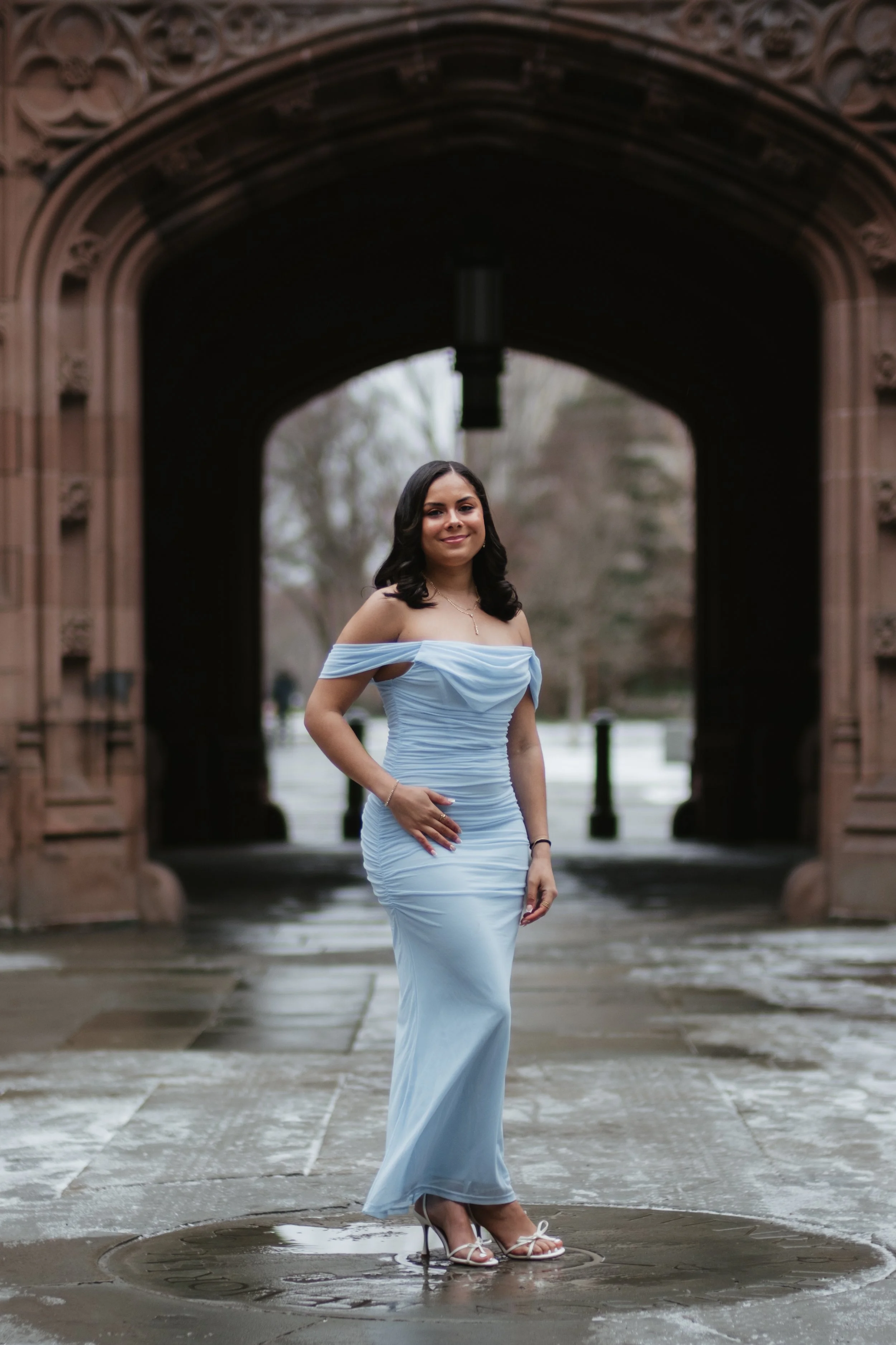 A young woman in a light blue off-shoulder evening gown and white heels stands outdoors in front of an ornate arched stone gateway, with trees and a paved walkway in the background.