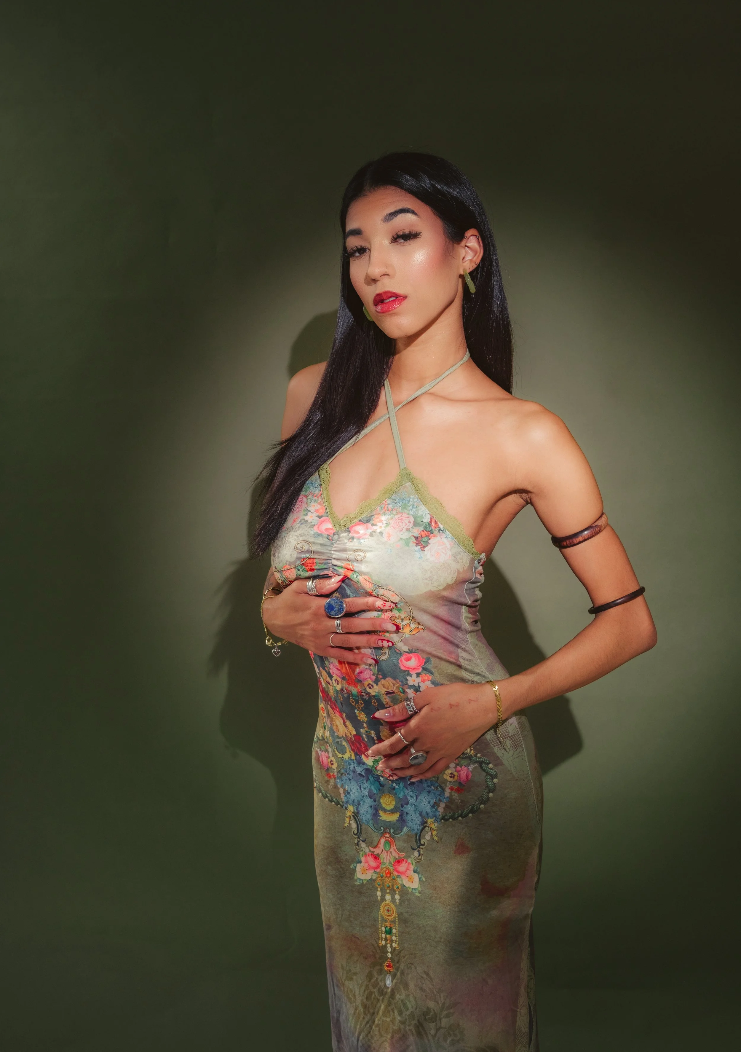 A woman with long black hair, wearing a floral dress with a greenish tint, standing against a dark green background.
